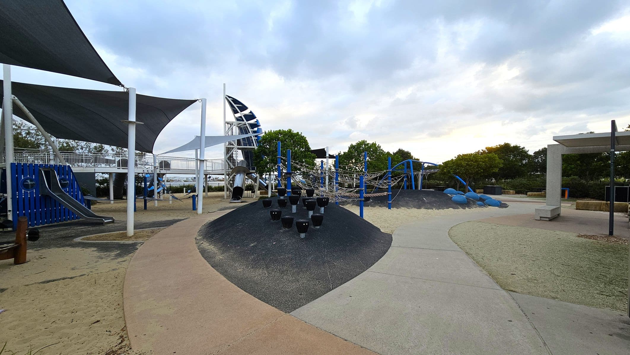 A playground with various equipment. On the left, there are blue and silver slides under large triangular shade sails supported by white poles. In the centre, a black mound with small climbing structures and a rope climbing frame with blue supports. On the right, a path leads to a shaded seating area. The ground is a mix of sand and rubber surfaces, and there are trees in the background.