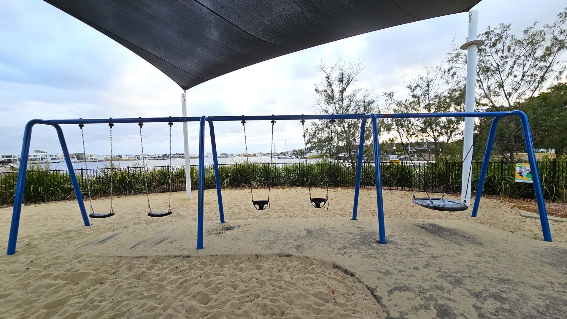 A playground with a blue swing set featuring four swings: two regular swings, two baby swings, and a circular net swing. The ground is covered in sand with patches of green matting. In the background, there is a body of water with boats and a line of modern buildings. Trees and shrubs are visible along a fence, and a large shade sail is overhead.