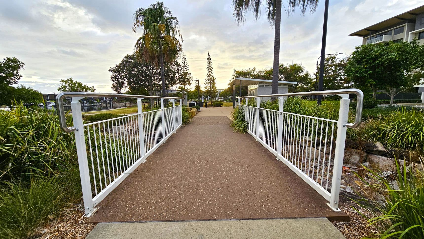 A pathway with a reddish-brown surface leads over a small bridge with white railings. On either side, there are green plants and grass. In the background, there are tall trees, including a palm tree on the left and a conifer in the centre. To the right, a modern building is partially visible. The sky is overcast with light clouds.