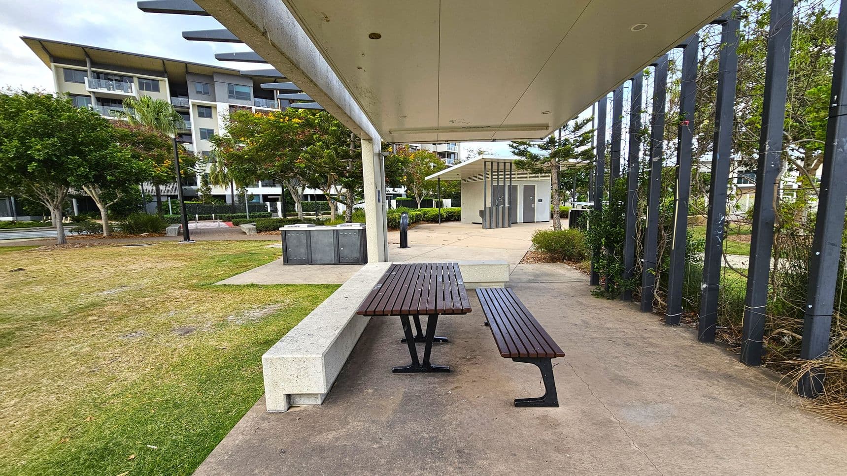A covered outdoor area with a wooden picnic table and bench on a concrete floor. To the right, vertical metal beams support climbing plants. In the background, a small building with doors is visible, surrounded by trees and bushes. On the left, a grassy area with more trees and a multi-storey building can be seen. The ceiling is flat and extends over the seating area.