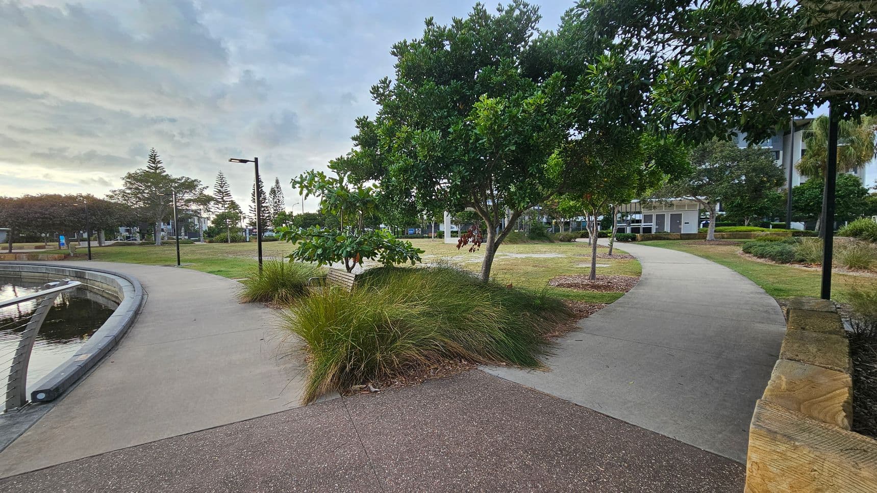 A park scene with two diverging concrete paths. The left path runs alongside a body of water with a low railing, while the right path curves through grassy areas and trees. In the centre, there are bushes and a bench beneath a tree. Several lamp posts line the paths. In the background, there are more trees and a building. The foreground features a reddish-brown textured surface and a wooden edge on the right.