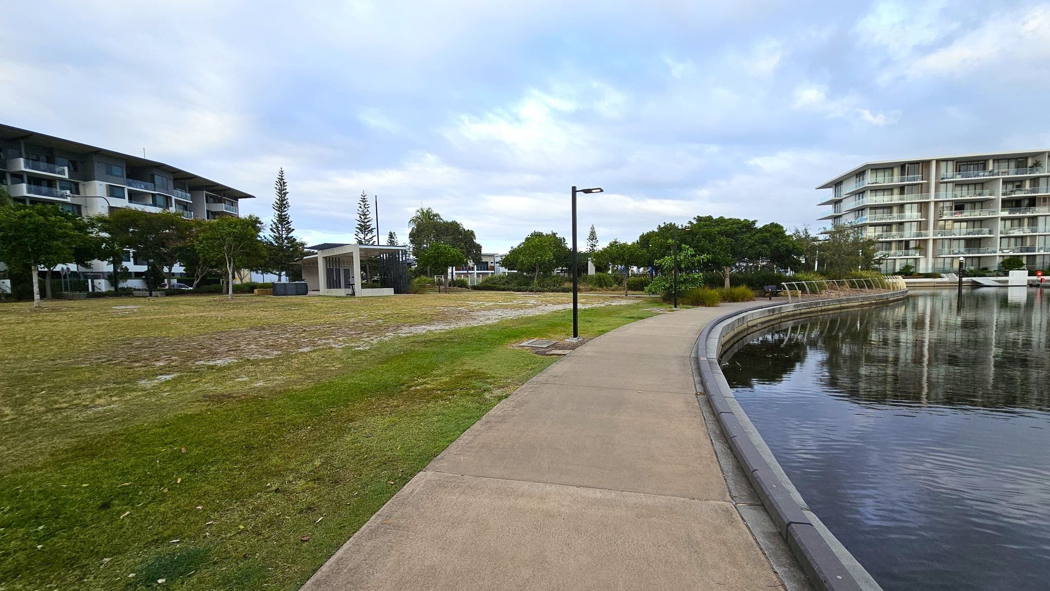 A curved pathway runs alongside a body of water on the right, with reflections of nearby buildings visible on the surface. On the left, there's a grassy area with scattered trees and a modern, open-sided shelter. Two multi-storey buildings are visible in the background, one on each side, with balconies and large windows. Street lamps line the path, and the area appears to be a landscaped urban park.