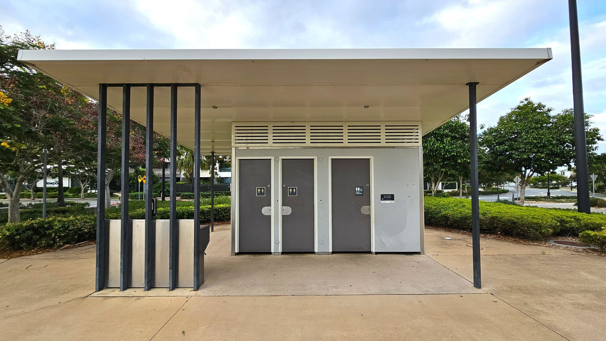 A public toilet block with a flat roof and three grey doors, each marked with gender-neutral signs. The structure has vertical black metal supports on the left. The ground is paved with light brown concrete. In the background, there are green bushes and trees with some red and yellow leaves. A road and a few parked cars are visible further back.