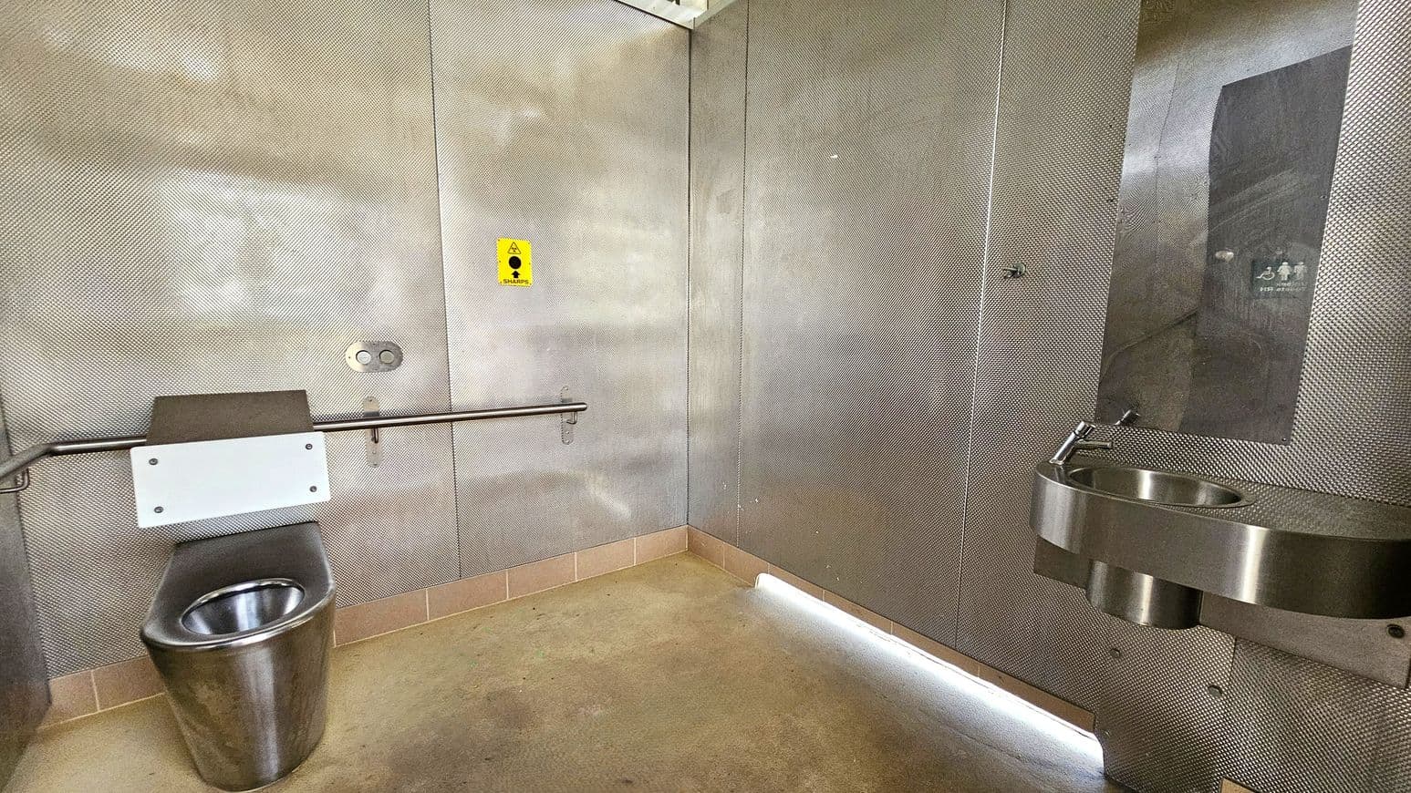 A public restroom with metallic walls and a concrete floor. On the left, there is a stainless steel toilet with a white panel and a horizontal grab bar. A yellow sharps disposal sign is visible above. On the right, a stainless steel sink is mounted on the wall with a small mirror above it. The ceiling is not visible.