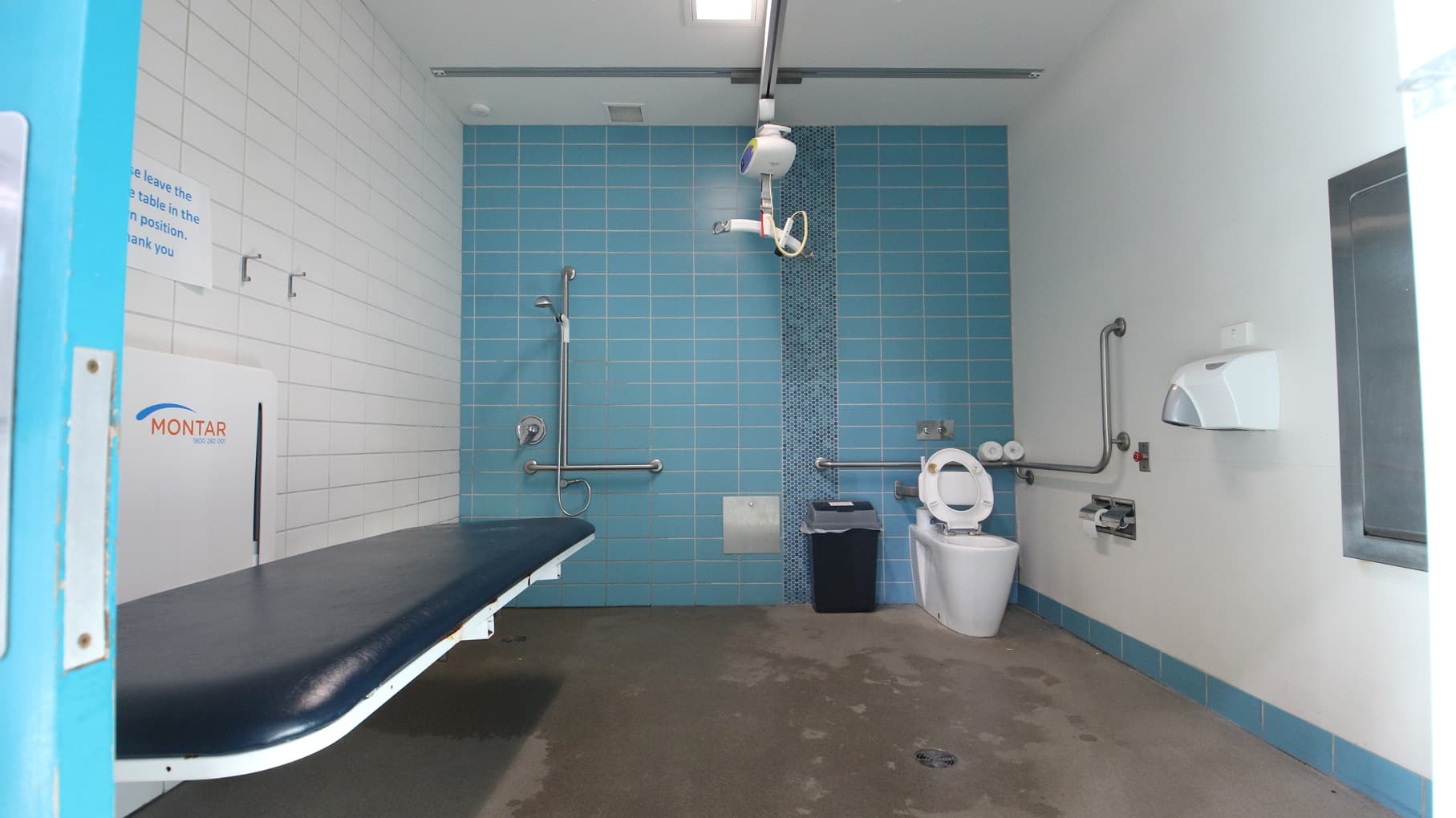 Inside the accessible bathrooms at Splash Devonport is a hoist, a dark blue cushioned adult change table, a toilet, a shower, and guard rails. Beside the toilet bowl is the rubbish bin. The back wall is made of blue tiles, the left is white tiles, while the right is a smooth white wall.