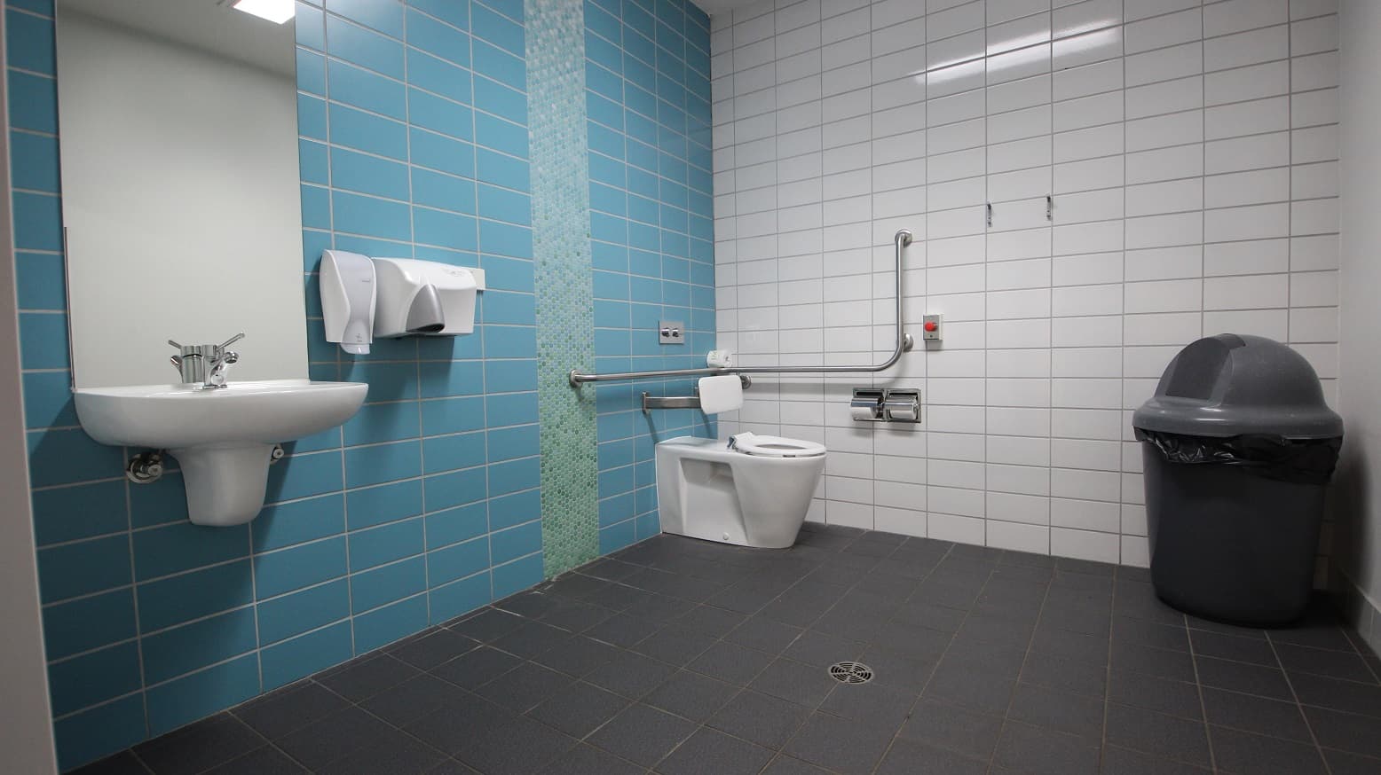 Another accessible bathroom at Splash Devonport. On the blue tiled wall are the wash basin, a large mirror, a liquid soap dispenser, and a hand dryer. This bathroom has a grey and black large rubbish bin placed opposite the toilet bowl with a white cushion to support the back of guests. Metal handles for tissue are mounted on the white tiled wall.