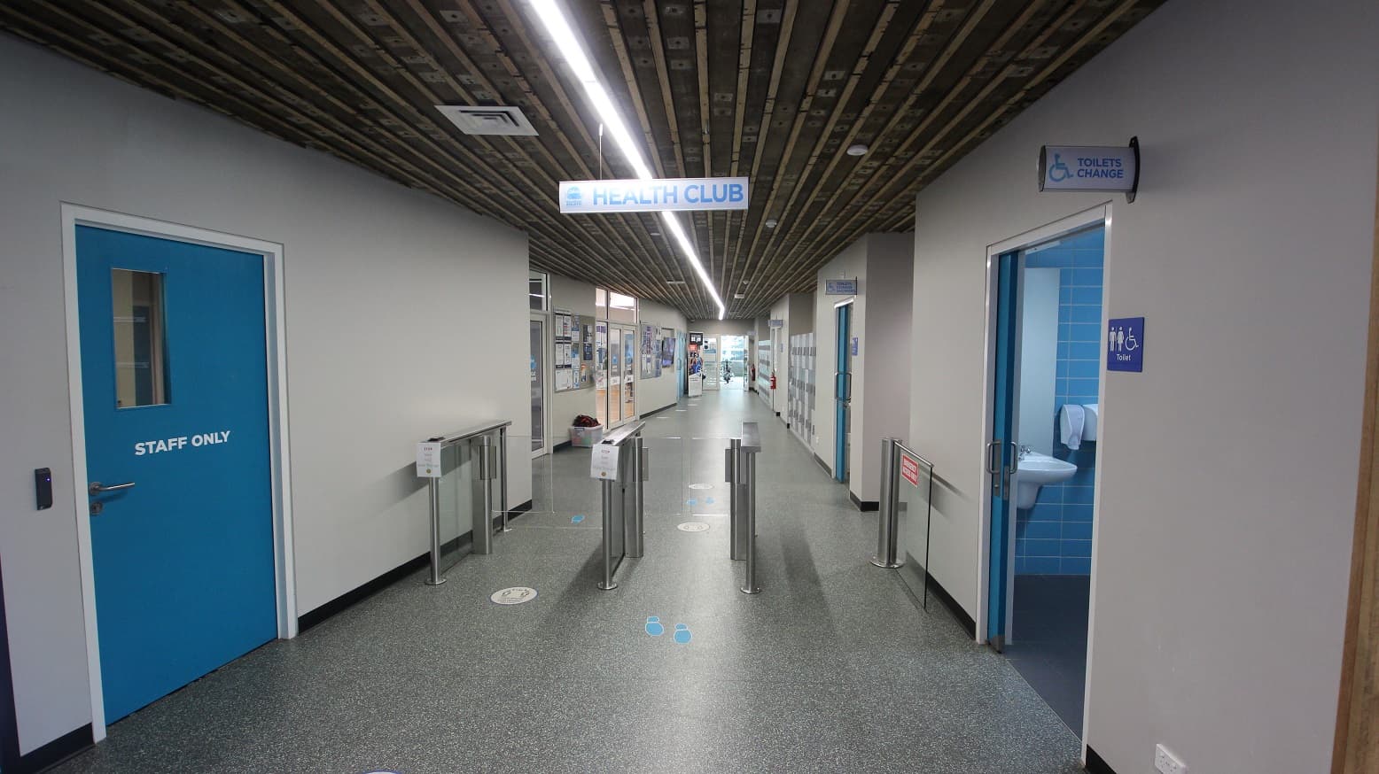 Turnstiles at the Health Club section at Splash Devonport. On the left side is the blue door with a metal handle with a staff-only label. To the right, is the Unisex Toilet Change area, which is designated by symbols for men, women, and people with mobility disabilities.
