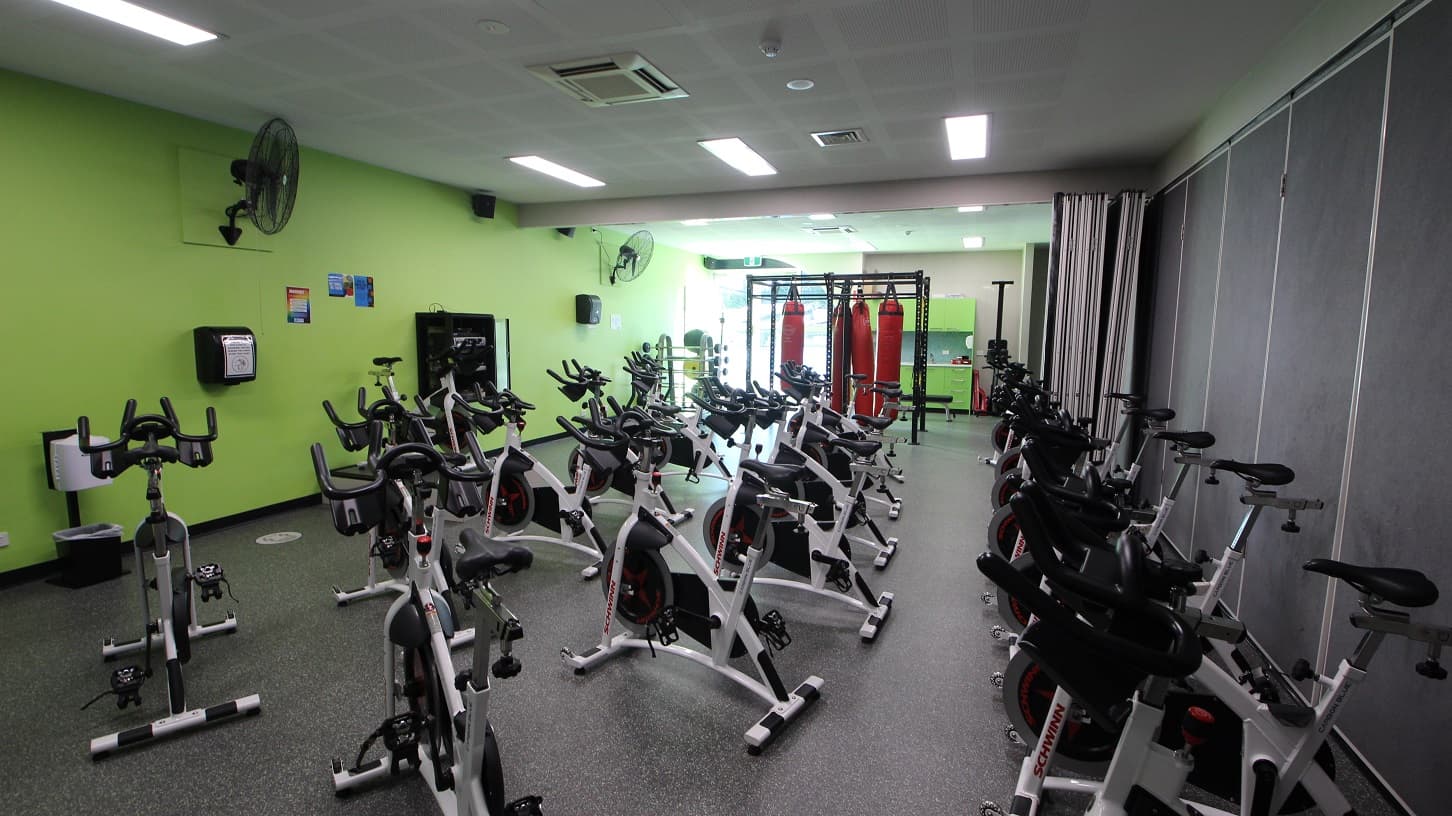Inside the cycle room at Splash Devonport. Several stationary bikes of grey, black, and white are arranged in three rows and are available for use. Movable walls painted in grey at the back separate the cycle room from the other. The other walls are painted neon green and a large, black, wall fans are mounted on the wall for ventilation. Four red punching bags are also hung on a black metal frame.