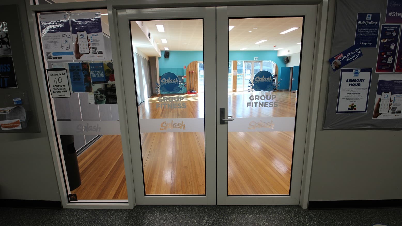 Manually operated glass double door of Group Fitness room at Splash Devonport. At the entrance door is a paper telling guests that only 40 people can utilize the facility at one time. Seen through the glass doors are the brown walnut wood flooring and the sky-blue wall and another glass door overlooking the outside pool. At the back of the room is the movable grey wall.