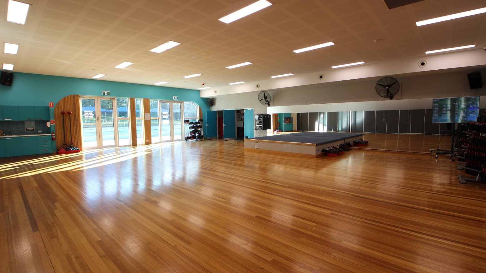 Large open space inside the Group Fitness gymnasium. The brown walnut wooden flooring shines beautifully as light strikes through it. Large mirrors are placed on the wall in front. A stage with a grey top that leaders could step into during fitness dance. Wall fans are mounted on the front wall of the room for proper ventilation and two blue doors on the left side are open.