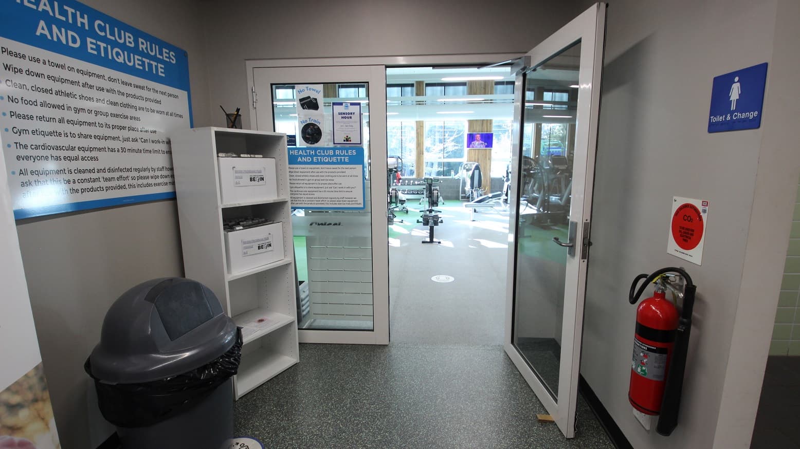 The other half of the manually operated double doors of Health Club at Splash Devonport is open while the other is not. Health Club rules and etiquette can be found on the doors and walls in small and large fonts for everyone to read. There is a large, grey, and black trash bin, a white cabinet, and a fire distinguisher at the entrance doors.