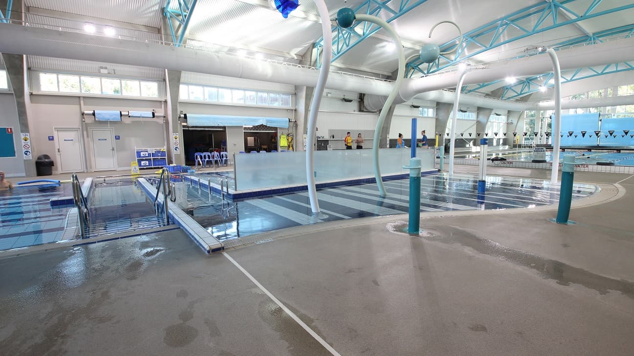 Automatic entrance doors at the Aquatic Centre in Splash Devonport. On the left side is the locker area with rectangular and square locker cabinets and their designated keys. The venue has very high ceilings with a corrugated iron finish and solid light blue steel archways. Large air conditioning white tubes dominate the ceiling space.