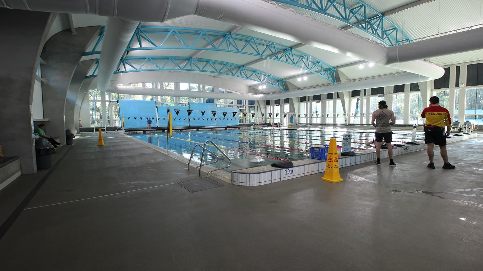 In 8 Lane 25m Pool is a hanging of small triangular flags placed horizontally to the pool area. The pool lanes are separated by red and yellow floating lane ropes, while the others are separated by red and blue lane ropes. Left of the pool is the stair, while on the right is the entrance ramp with a metal handrail. The starting point is an elevated section with different depth marks on each lane.