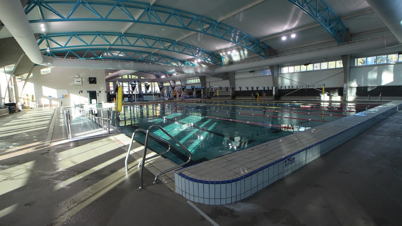 The 1.75-meter depth lane of the 25-meter pool in Splash Devonport aquatic centre. This side of the pool also has a staircase. Evident in this image are the high ceilings with a corrugated iron finish and solid light blue steel archways. Large air conditioning white tubes dominate the ceiling space.