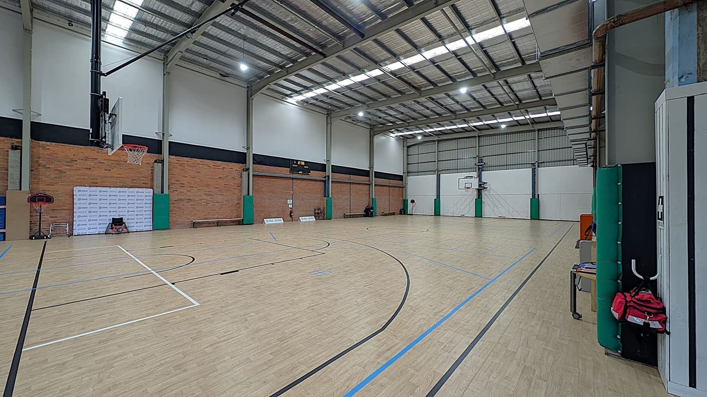 A reception area with a curved white desk in the centre, displaying brochures and a computer monitor. Behind the desk is a wall with vertical white slats. To the left, a black leather sofa and a framed sports jersey are visible. On the right, a green banner advertising exercise therapy stands near gym equipment. The floor is dark with a speckled pattern, and the ceiling is white with recessed lighting.
