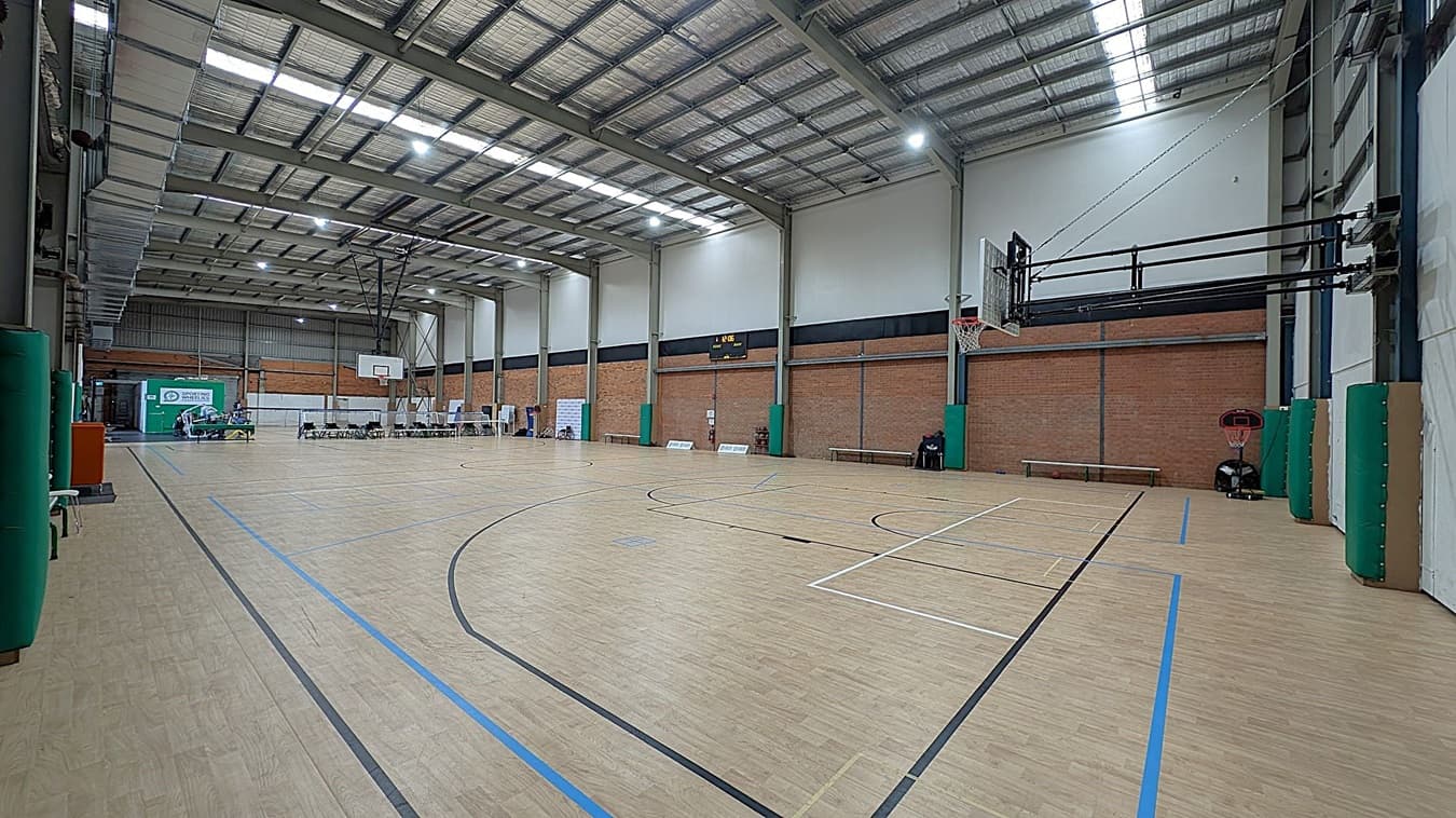A spacious indoor basketball court with a light wooden floor marked with black and blue lines. On the right, a basketball hoop is mounted on the wall. The left side features a row of chairs and tables near a green and white sign. The walls are a mix of brick and white, with green padding on the columns. The ceiling is high with exposed metal beams and bright lights. A scoreboard is visible on the far wall.
