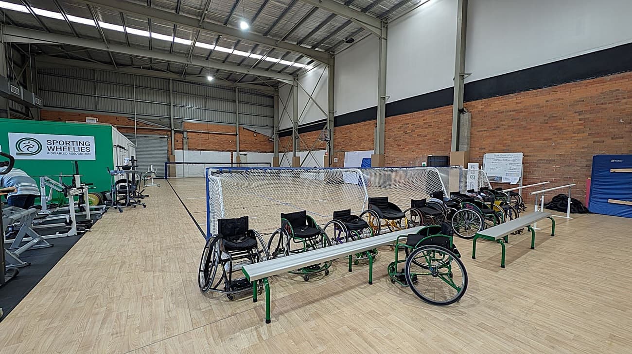 A sports hall with a wooden floor and high metal ceiling. On the right, there are several wheelchairs lined up behind a bench. In the centre, a net separates the area. On the left, exercise equipment is visible near a green wall with a "Sporting Wheelies & Disabled Association" sign. The background features brick walls and a whiteboard.