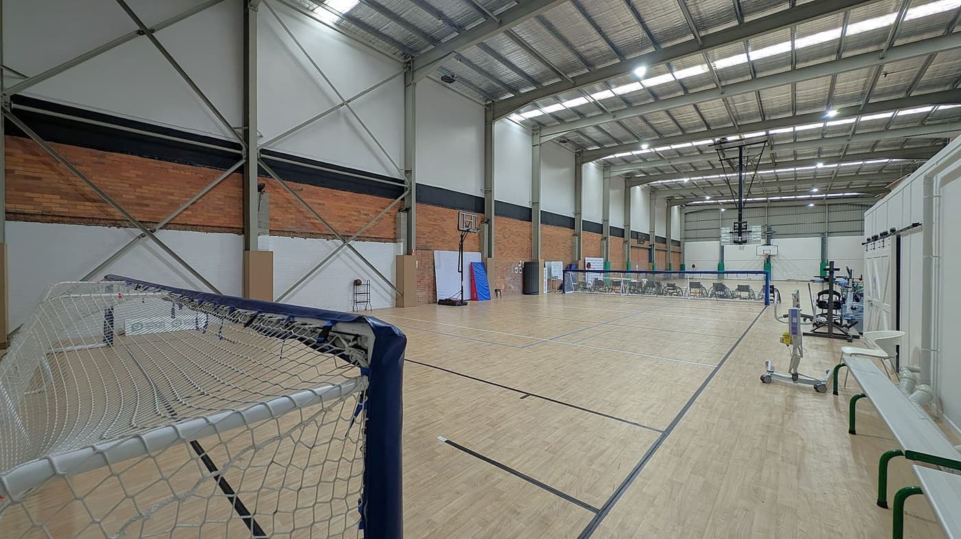 A spacious indoor sports hall with a wooden floor and high metal ceiling. On the left, a goal net is visible. The walls are a mix of brick and white panels with metal supports. In the centre, a basketball hoop and a badminton net are set up. On the right, there are gym equipment, benches, and chairs lined up along the wall. The ceiling has large fluorescent lights.