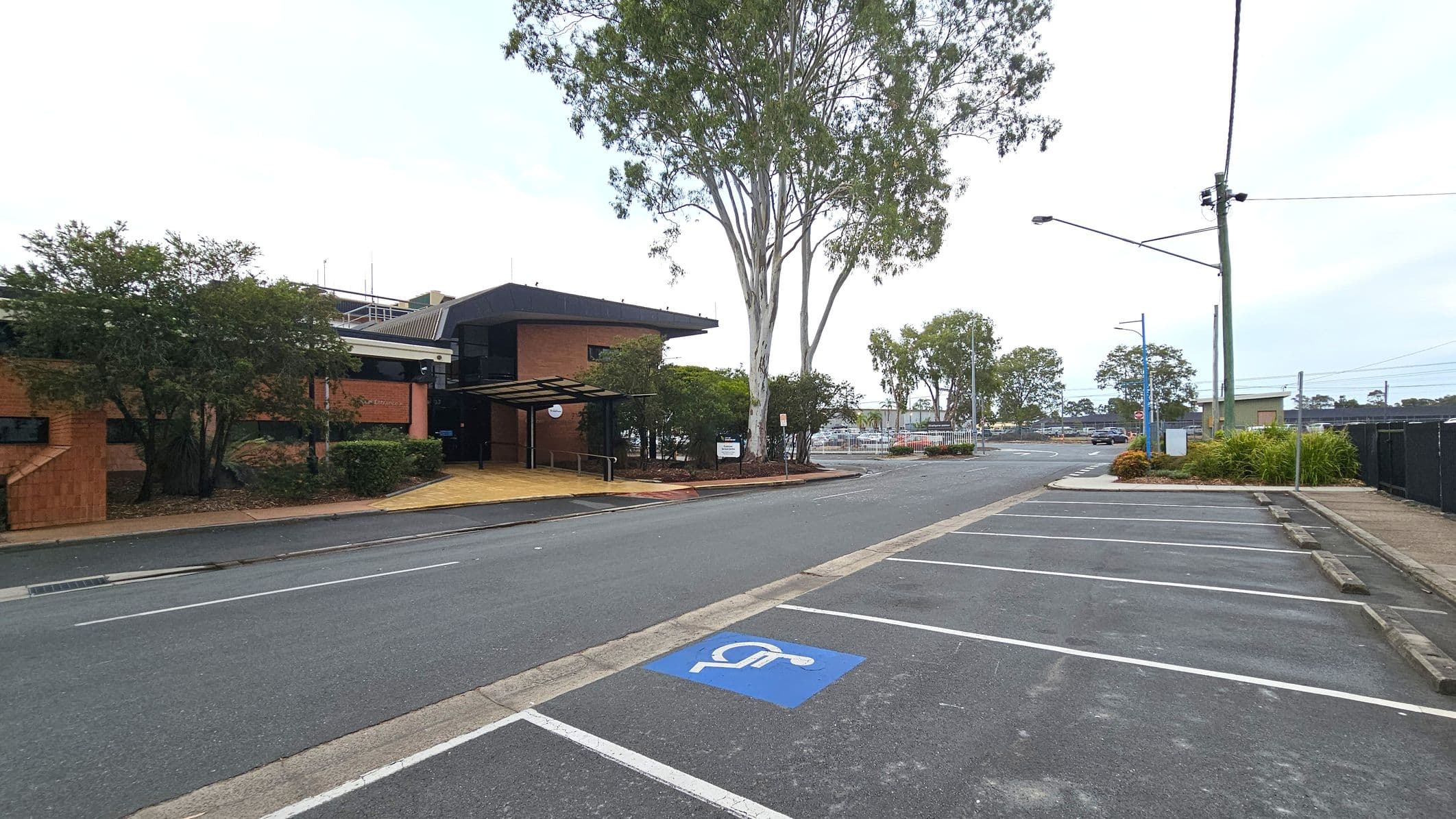 A street scene with a brick building on the left, featuring a covered entrance and surrounded by trees and shrubs. A large tree stands near the building. On the right, there is a parking area with marked spaces, including a designated disabled parking spot. The background shows more trees, a fence, and a few buildings. A streetlight and utility pole are visible near the parking area. The road is empty, and the pavement is bordered by a narrow footpath.
