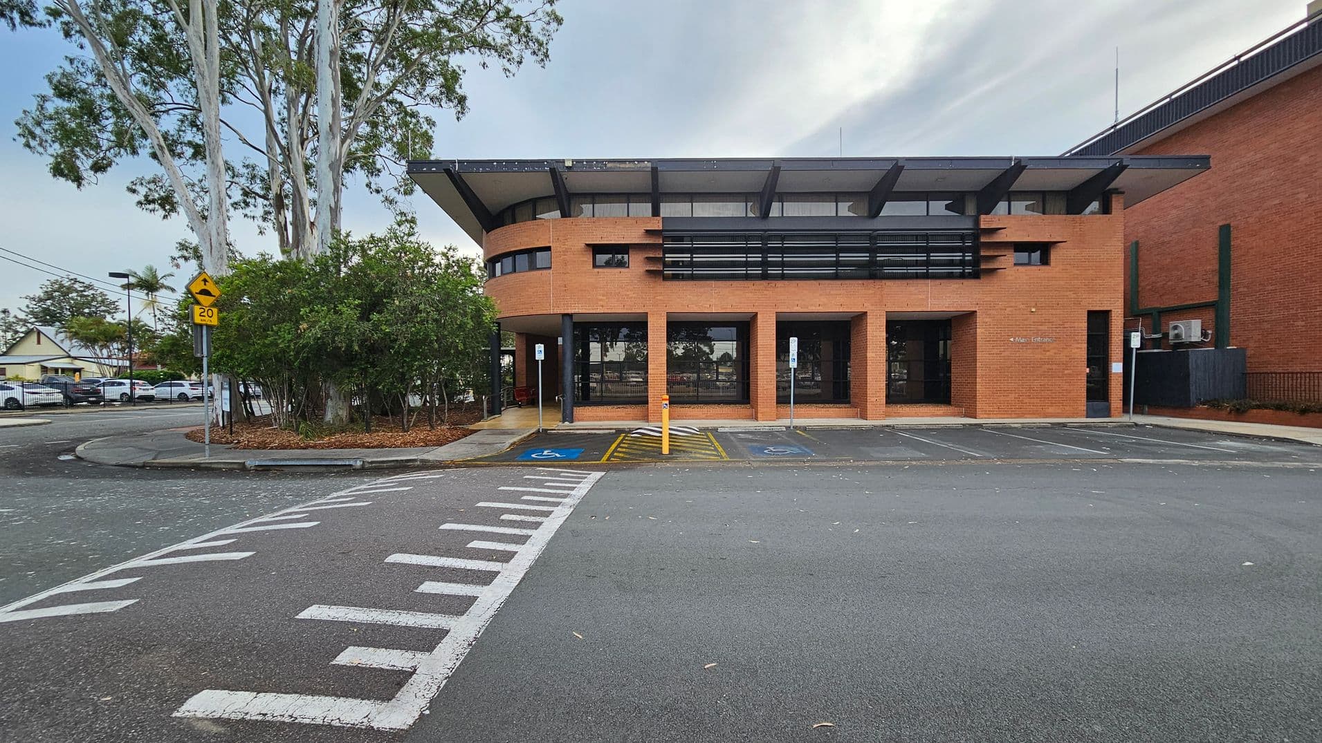 A brick building with a modern design features large windows and a flat roof with overhangs. In front, there are two marked disabled parking spaces and a pedestrian crossing. To the left, a cluster of trees and shrubs is visible, along with a yellow road sign indicating a speed limit of 20. The road curves around the building, and a few parked cars are seen in the background. The pavement is grey with white markings.