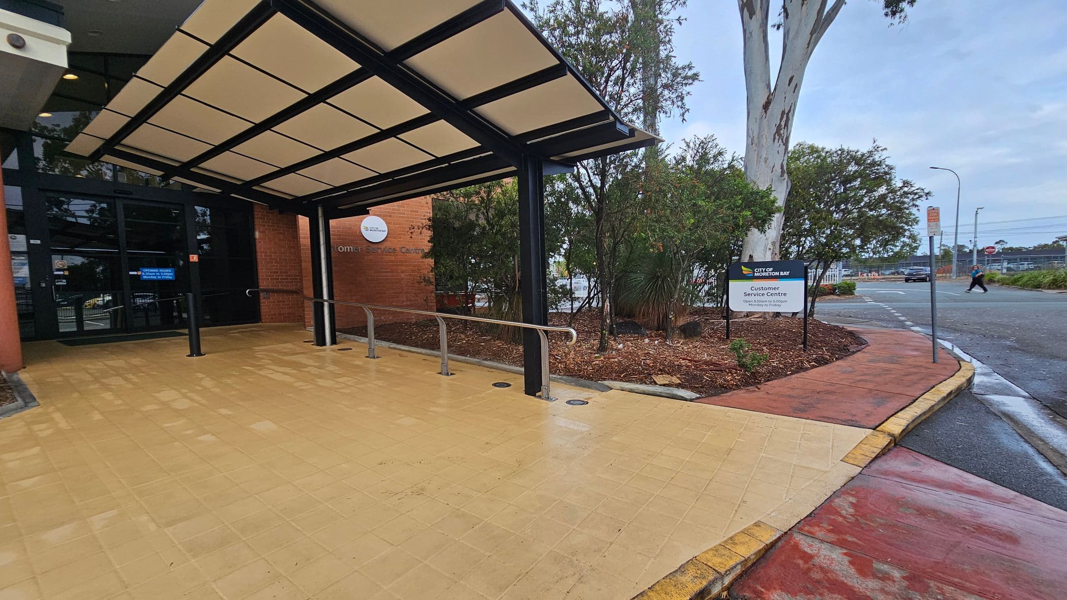 Entrance to a building with a covered walkway on the left, featuring a glass door and a sign indicating a customer service centre. The floor is tiled in a light yellow colour. On the right, there is a landscaped area with bushes and a tree, alongside a sign for the City of Kwinana. A pavement leads to a road where a person is crossing. The ceiling of the walkway is structured with a black frame and light panels.