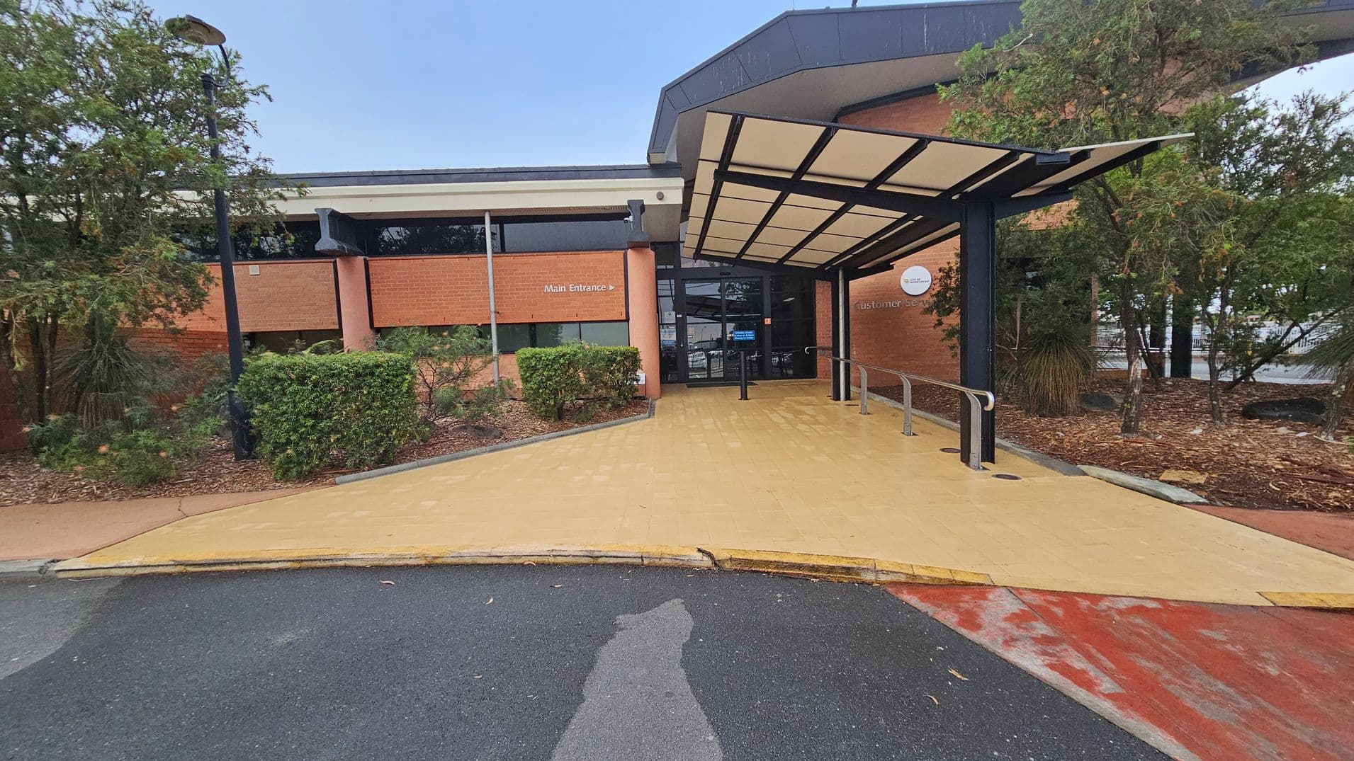 A building entrance with a covered walkway leading to glass doors. The structure has a brick facade with a sign indicating the main entrance. On the left, there are bushes and a lamp post, while on the right, more trees and shrubs are visible. The ground is paved with a yellowish surface leading from the road, which is partially visible in the foreground. The roof of the entrance is supported by black metal beams.