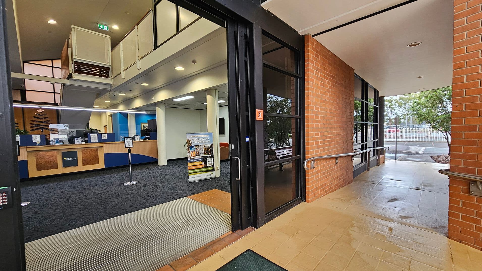 Entrance to a building with a glass door on the right, leading to a tiled corridor with brick walls and metal handrails. Inside, a reception area with a wooden counter and blue accents is visible. A sign with information stands on the carpeted floor. The ceiling has recessed lighting, and a staircase is visible in the background. Outside, trees and a parking area can be seen through the glass.