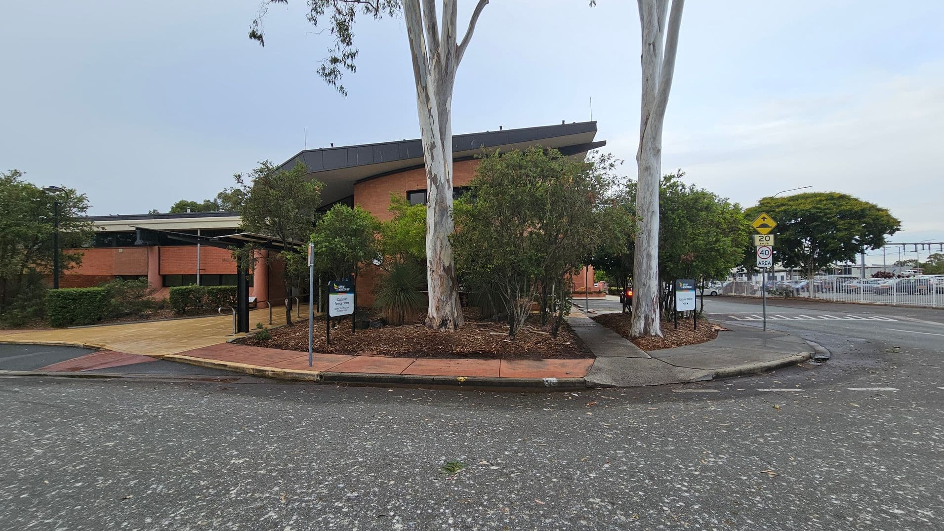 A brick building with a sloped roof is in the centre, surrounded by trees and shrubs. A large tree stands prominently in front. On the right, a road with speed limit signs curves around the building. A footpath runs alongside the road, bordered by landscaped garden beds. In the background, a fence and parked cars are visible. The ground is paved with a mix of asphalt and concrete.