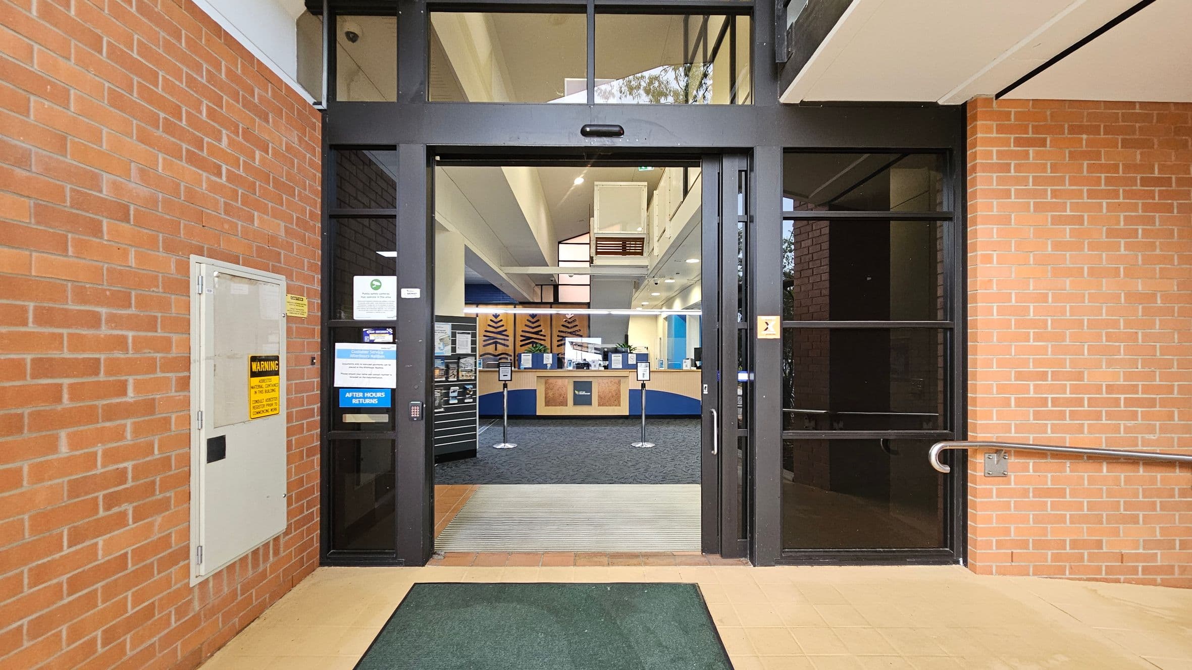 Entrance to a building with a glass door framed in black, flanked by red brick walls. On the left, a white panel with warning signs is mounted on the brick wall. Inside, a reception area with a counter and signage is visible. The floor outside is tiled with a green mat, and inside, the floor is carpeted. The ceiling is high with recessed lighting. A metal handrail is attached to the right wall.
