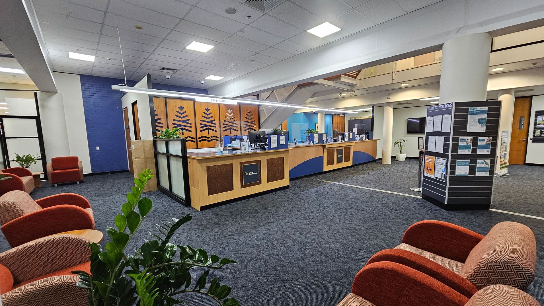 A spacious interior with a reception desk in the centre, featuring wood panelling and decorative designs. To the left, there are several orange upholstered chairs and green plants. On the right, a board displays public notices. The floor is carpeted in a dark blue pattern, and the ceiling is white with recessed lighting. In the background, there are glass doors and a staircase leading to an upper level.