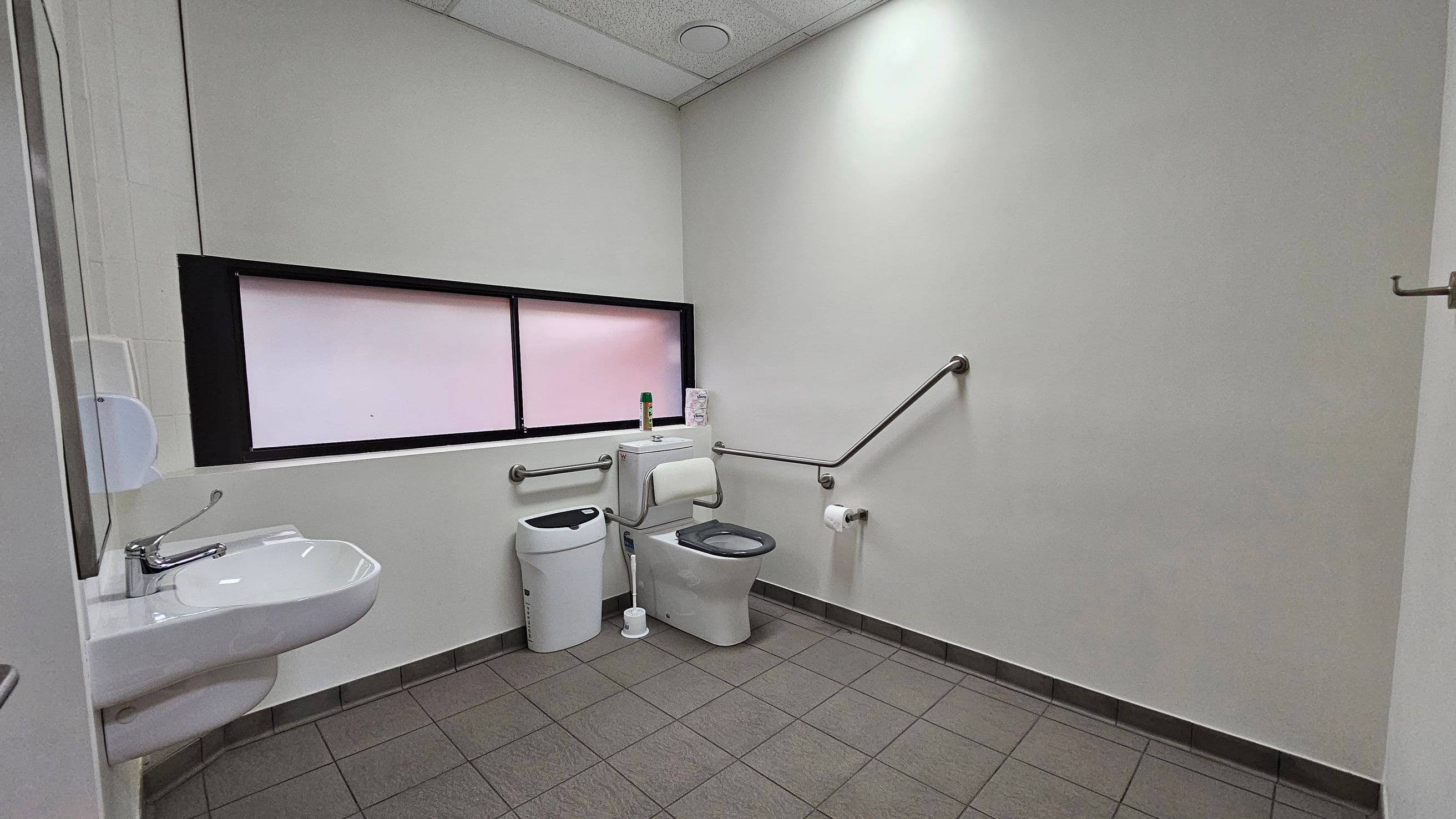 A public restroom with grey tiled flooring and a white ceiling. On the left, a white sink with a silver tap is mounted on the wall, accompanied by a soap dispenser. In the centre, a toilet with a grey seat is positioned next to a sanitary bin and a toilet brush. Above the toilet, a window with frosted glass is visible. Grab bars are installed on the walls near the toilet, and a toilet roll holder is mounted on the right wall.