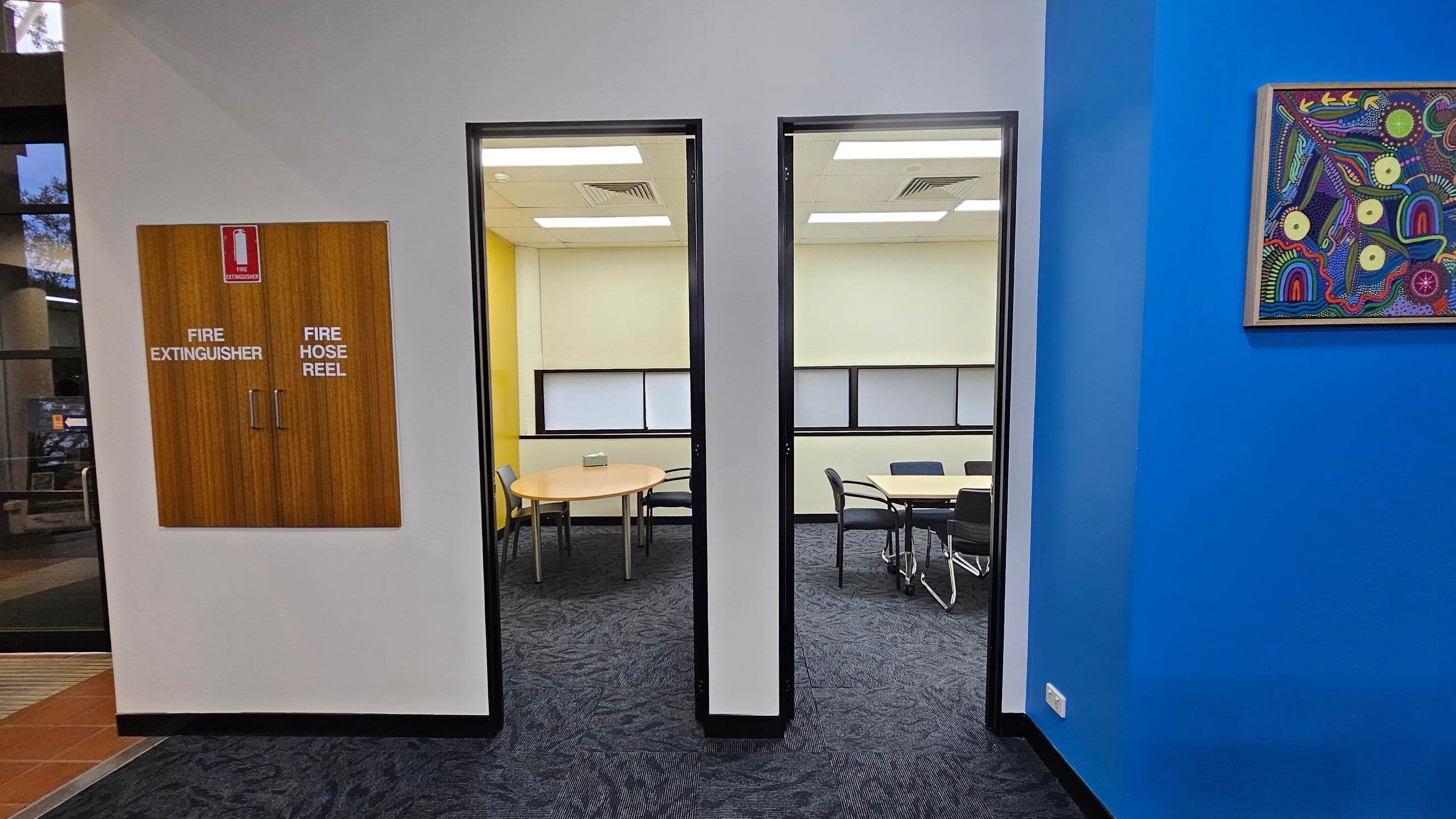 A hallway with a grey wall in the centre featuring a wooden cabinet labelled "Fire Extinguisher" and "Fire Hose Reel". Two doorways lead to rooms with tables and chairs. The left room has a yellow wall, while the right room has a light-coloured wall. On the right, a bright blue wall displays a colourful abstract painting. The floor is carpeted in a dark pattern, and the ceiling has white panels with recessed lighting.