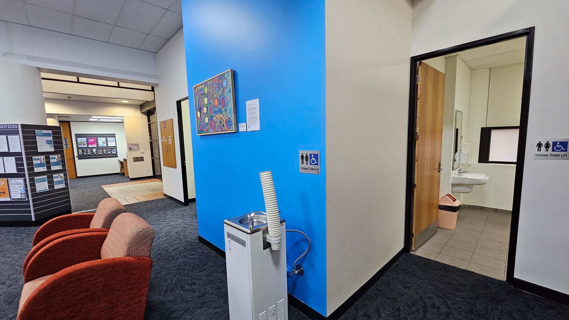 A hallway with a blue accent wall in the centre, featuring a colourful artwork. Below the artwork is a water fountain with stacked cups. To the right, an open door leads to a unisex accessible toilet, visible with a sink and bin. On the left, two red chairs are placed on a dark carpeted floor. Further left, a notice board displays various papers. In the background, a corridor with a light-coloured floor and a wooden door is visible. The ceiling is tiled.