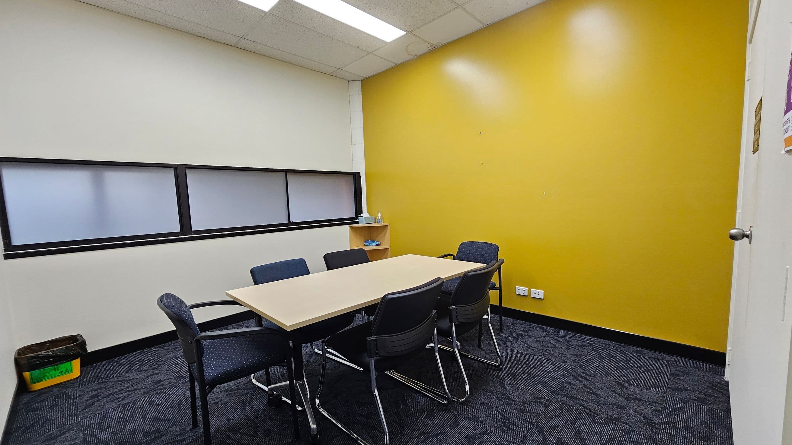A small meeting room with a rectangular table surrounded by six chairs. The left wall is white with a horizontal frosted window, and a yellow bin is on the floor. The right wall is bright yellow with a small shelf holding a bottle and some items. The floor is covered with dark patterned carpet tiles. The ceiling has white panels with fluorescent lights.