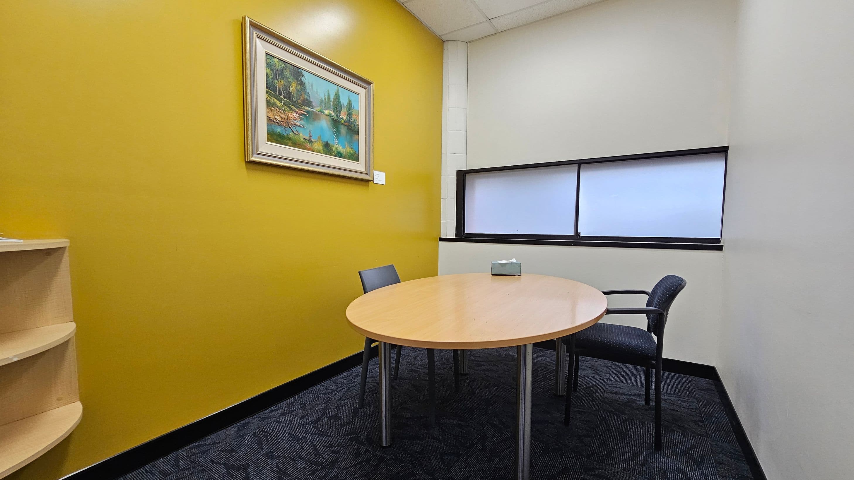 A small office room with a mustard yellow wall on the left, featuring a framed landscape painting. Below the painting is a light wood shelf. In the centre, a round wooden table with a tissue box on top is surrounded by two chairs. The right wall is white with a rectangular window featuring frosted glass. The floor is covered with dark patterned carpet, and the ceiling is white with recessed lighting.