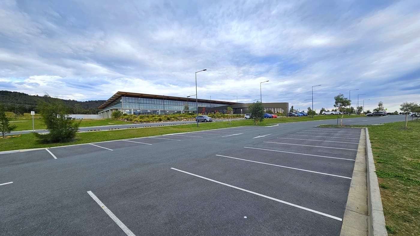 The Stromlo Leisure Centre has a contemporary architectural design incorporating glass, steel, and timber elements. Adjacent to the building, there is ample car parking with a few cars parked. Tall street lights, along with trees and grass, surround the area.