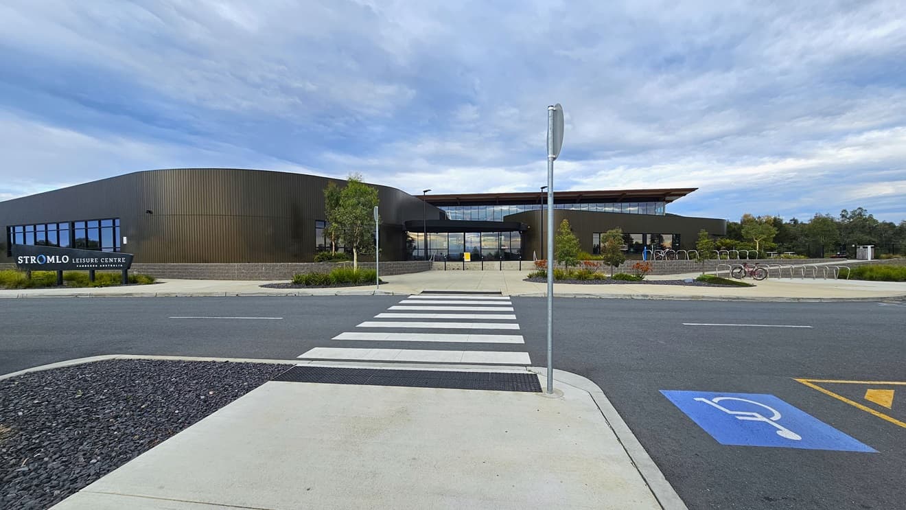The Stromlo Leisure Centre has a modern architectural design, blending glass, steel, and timber elements. A facility name sign stands in front of the building on the left. On the right, bike racks are available, and adjacent accessible parking is marked with blue wheelchair signs. A pedestrian lane in the middle is marked with truncated domes. A gravel area is situated on the left. Tall street lights, trees, and grass surround the facility.