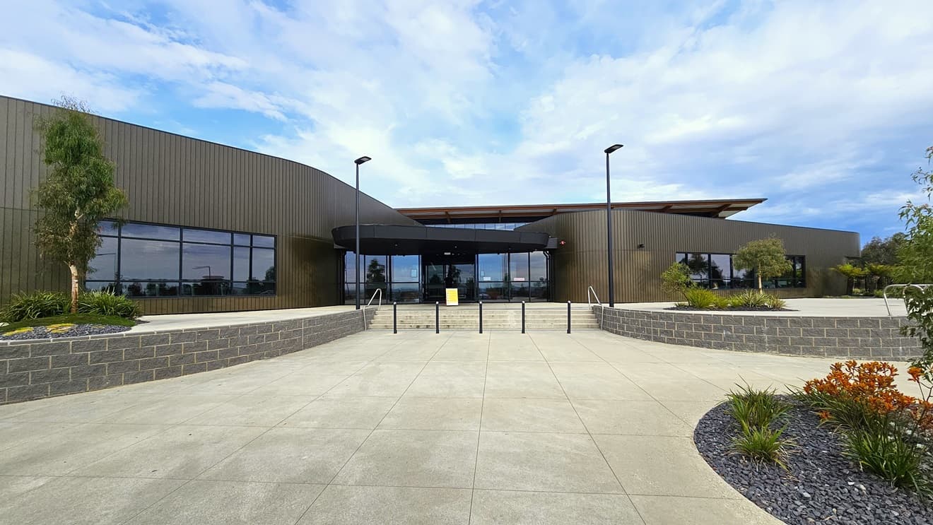 The Stromlo Leisure Centre features a contemporary architectural design, integrating glass, steel, and timber elements. A concrete staircase with handrails and bollards is positioned in the middle. The concourse is adorned with a landscape of plants, trees, and grass. Tall street lights are situated in front of the entrance.