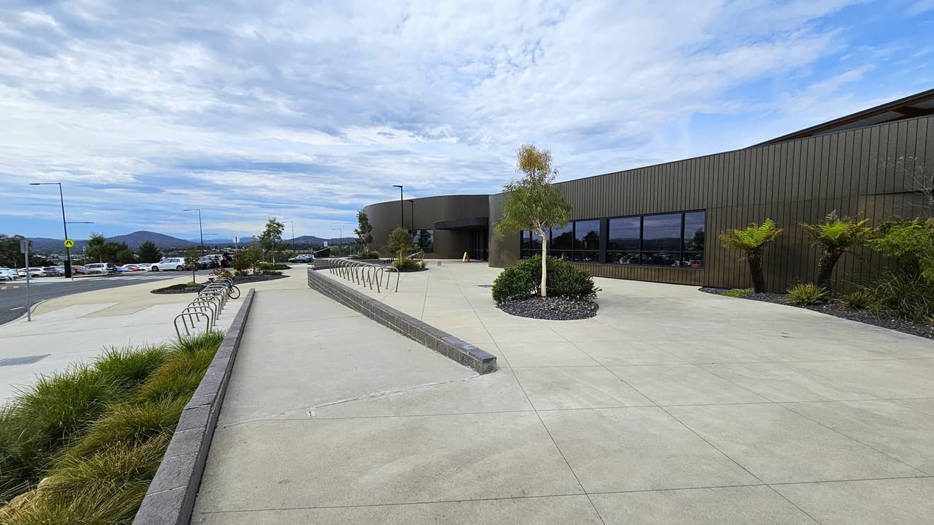 The Stromlo Leisure Centre has a contemporary architectural design, blending glass, steel, and timber elements. A ramp entry is located on the left side of the entrance, accompanied by visible bike racks. The expansive concourse is enhanced with a landscape of plants, palm trees, and grass.