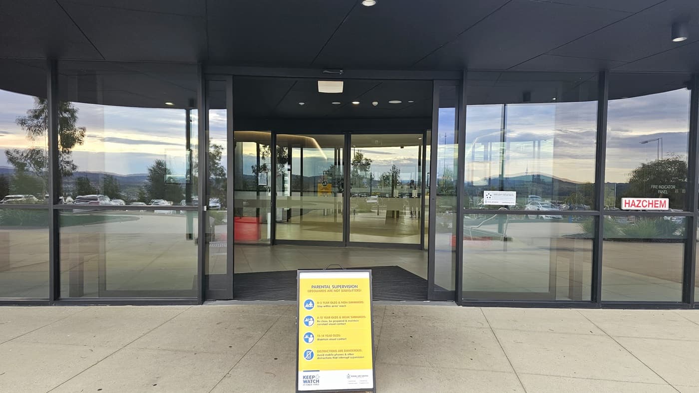The building entrance features two sets of automatic glass doors with non-slippery mats. A free-standing yellow banner is positioned in front of the entrance. Signs and safety stickers are affixed to the glass wall.