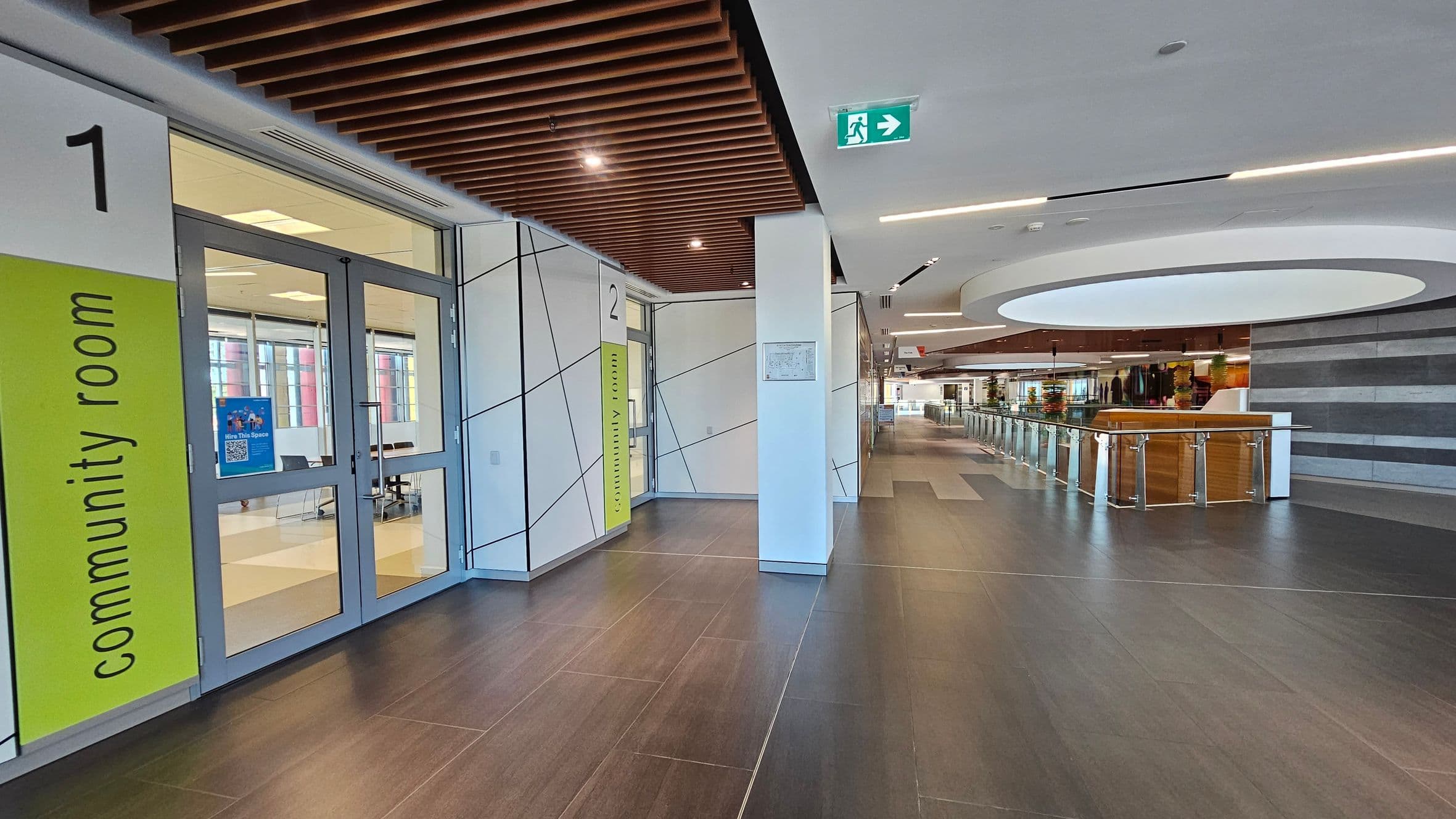A modern interior space with a dark tiled floor and a ceiling featuring wooden slats. On the left, there are two community rooms with glass doors, labelled "1" and "2" with green signs. In the centre, a corridor leads to a larger open area with a circular ceiling feature. On the right, there is a railing with glass panels and a view of a distant area with more seating and decor. An exit sign is visible on the ceiling.