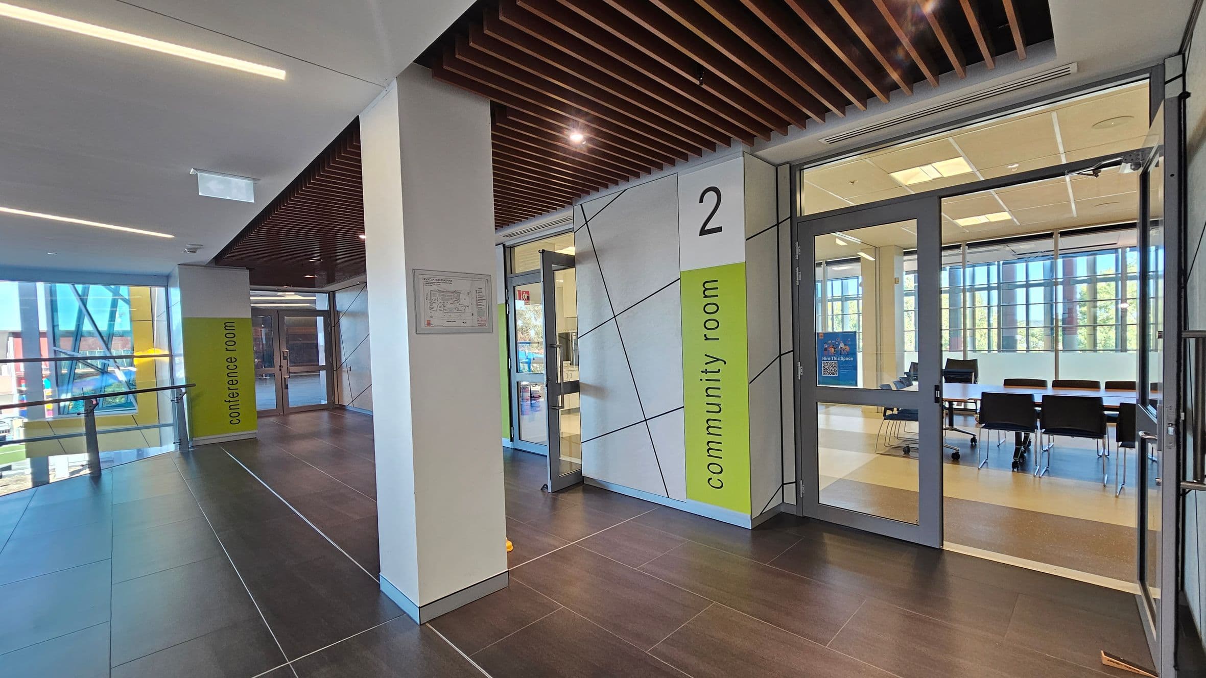 A modern interior space with dark tiled flooring and a ceiling featuring wooden slats. On the right, there is a glass entrance to a "community room" with visible chairs and tables inside. The wall next to the entrance is marked with the number "2" and labelled "community room" in green. On the left, a corridor leads to a "conference room" with large windows allowing natural light. The area is well-lit with recessed lighting.