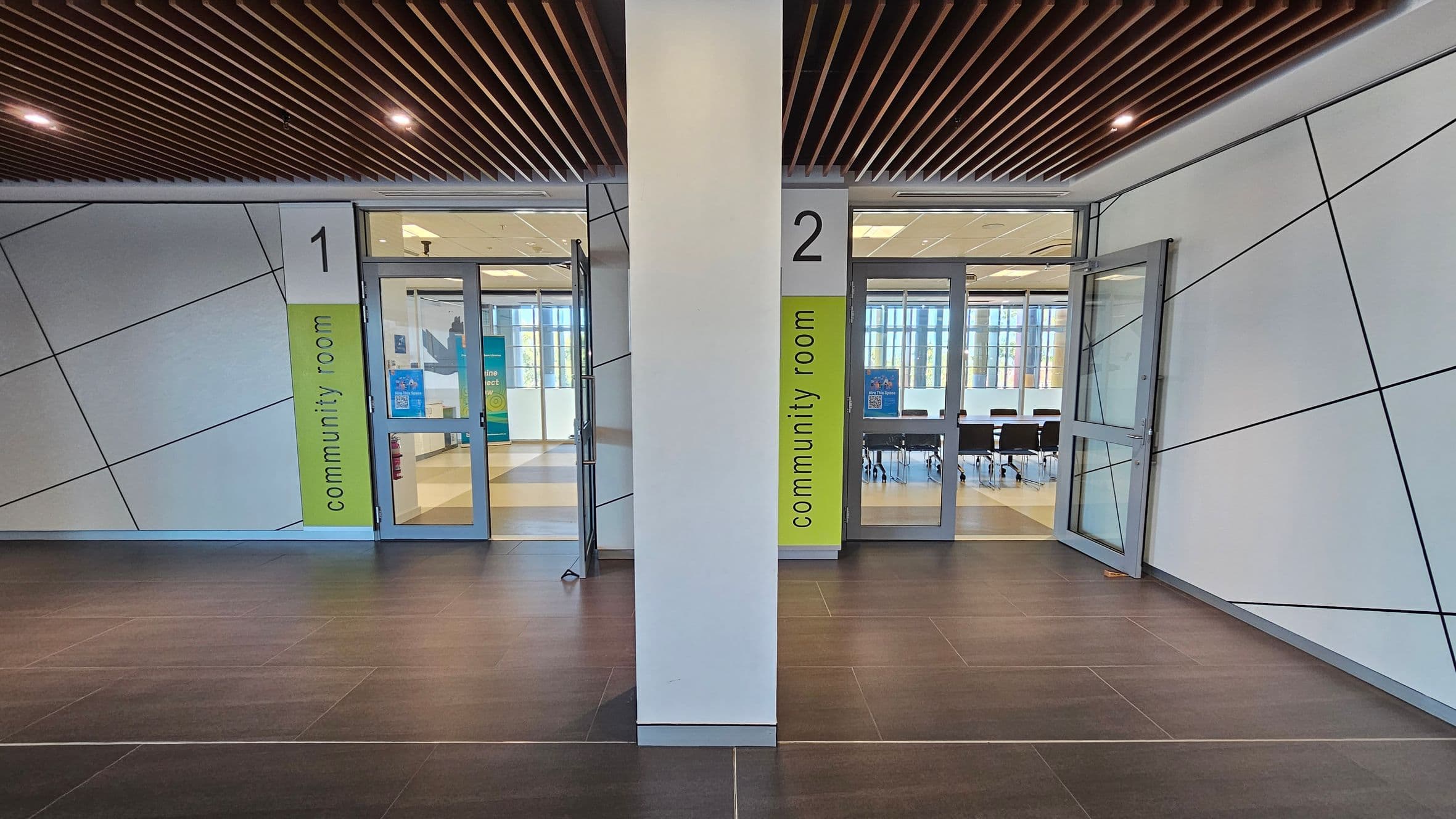A hallway with two community rooms, numbered 1 and 2, on either side of a central pillar. Each room has a glass door with a green sign indicating "community room." The walls have a geometric pattern with diagonal lines. The floor is dark tiled, and the ceiling features wooden slats with recessed lighting. Inside the rooms, tables and chairs are visible, along with large windows allowing natural light.