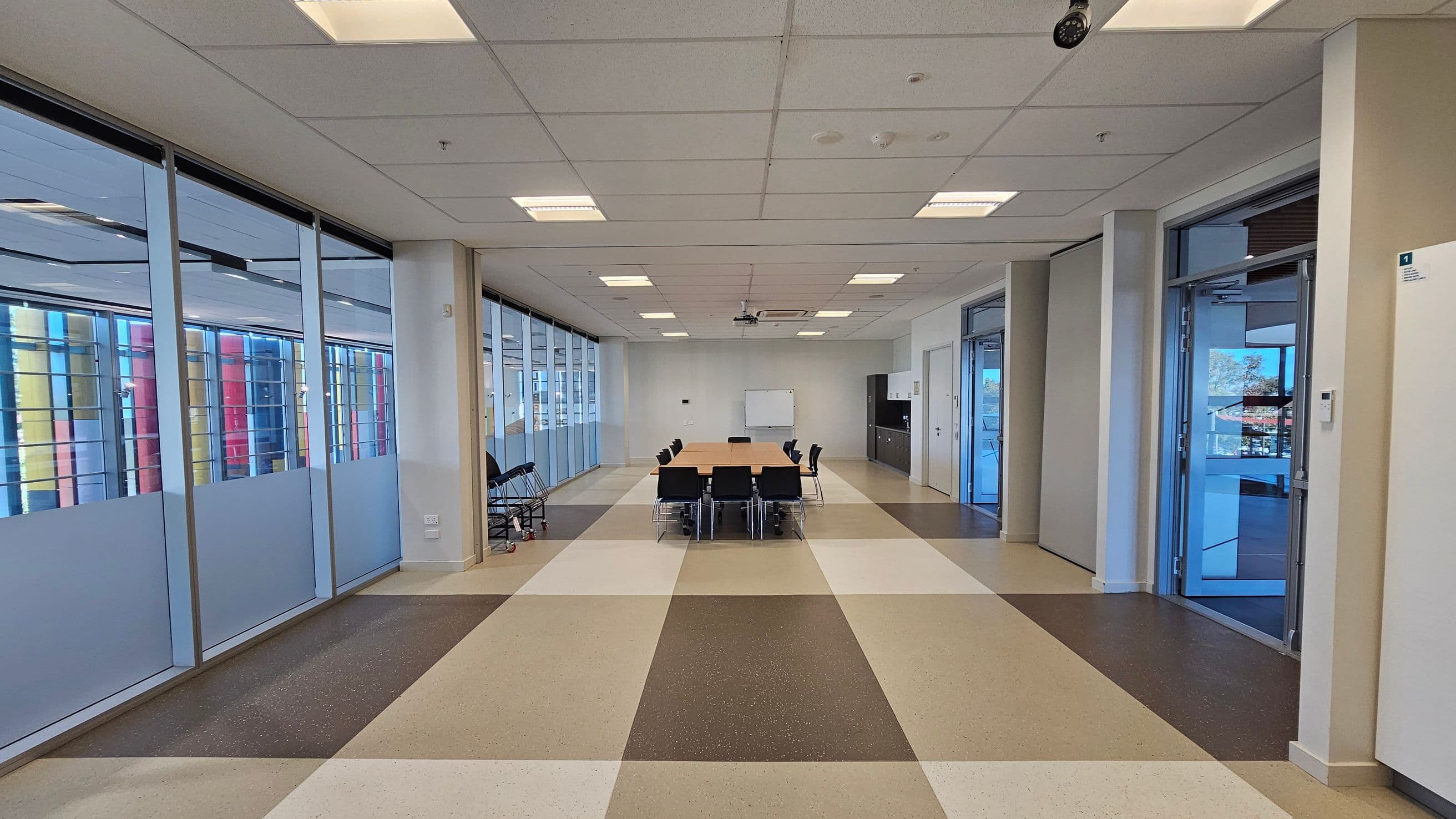 A spacious room with a long table and several black chairs in the centre. The floor has a pattern of large, alternating brown and beige tiles. On the left, there are large glass windows with frosted lower sections, showing colourful vertical structures outside. On the right, there are glass doors leading to another area. The ceiling is fitted with square lights, and a whiteboard is visible at the far end of the room.