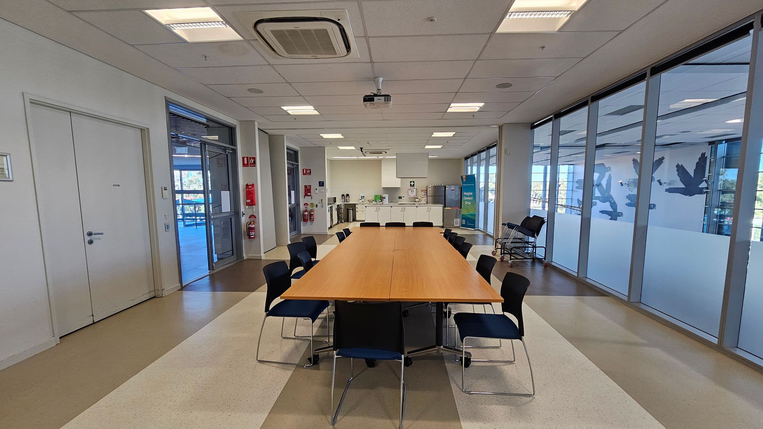 A conference room with a long wooden table in the centre, surrounded by black chairs. On the left, there are white doors and a glass exit door leading outside. The right side features large windows with frosted designs and a view of the outdoors. In the background, there's a kitchenette with appliances and cabinets. The ceiling has recessed lighting and an air conditioning unit. The floor is a mix of beige and brown tiles.