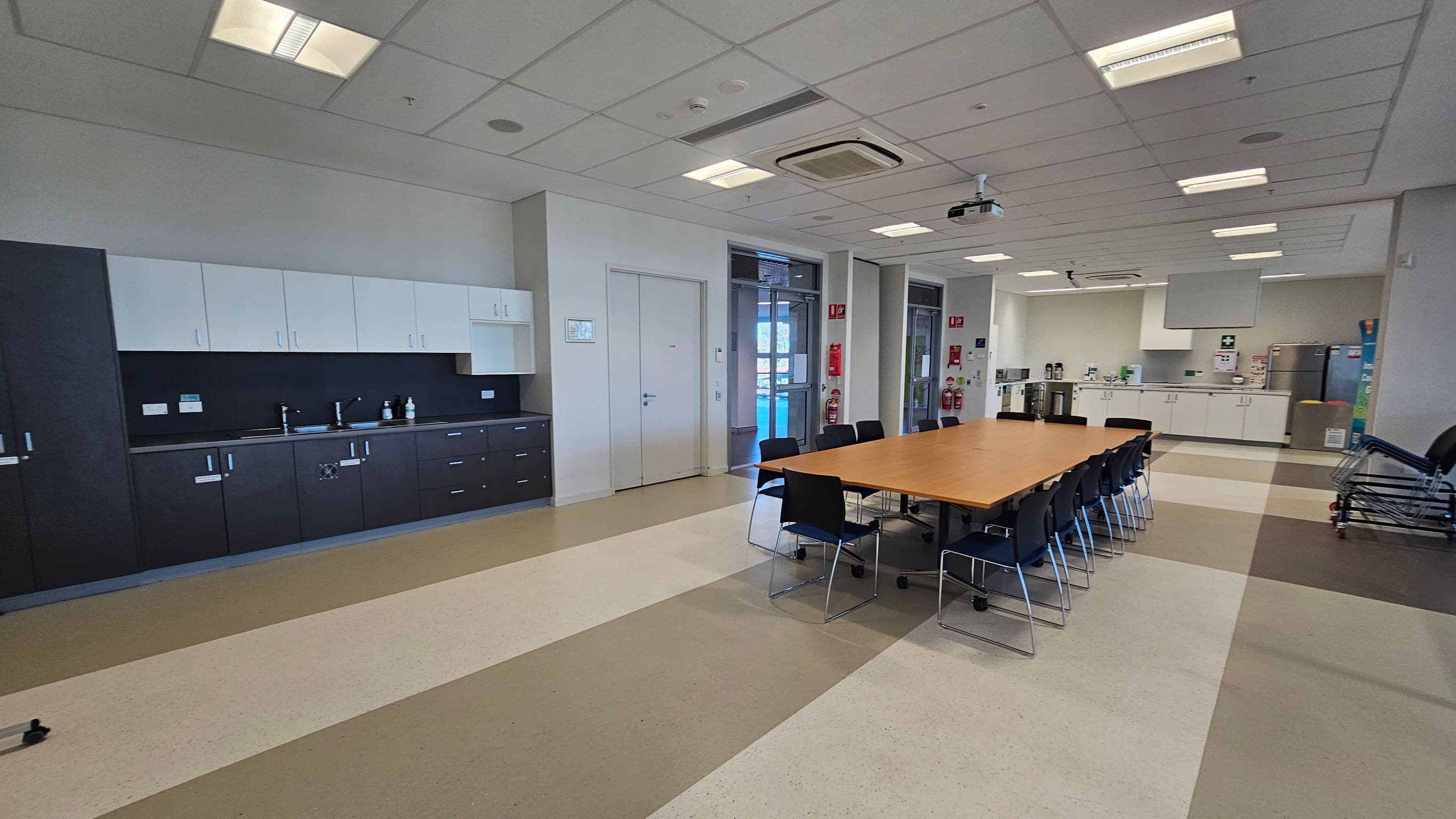 A spacious room with a long wooden table in the centre surrounded by black chairs. On the left, there is a wall with dark and light cabinets and a sink. The right side features a kitchenette with appliances and more cabinets. The floor has a pattern of beige and brown tiles, and the ceiling is fitted with fluorescent lights and an air conditioning unit. In the background, there are doors and fire safety equipment.