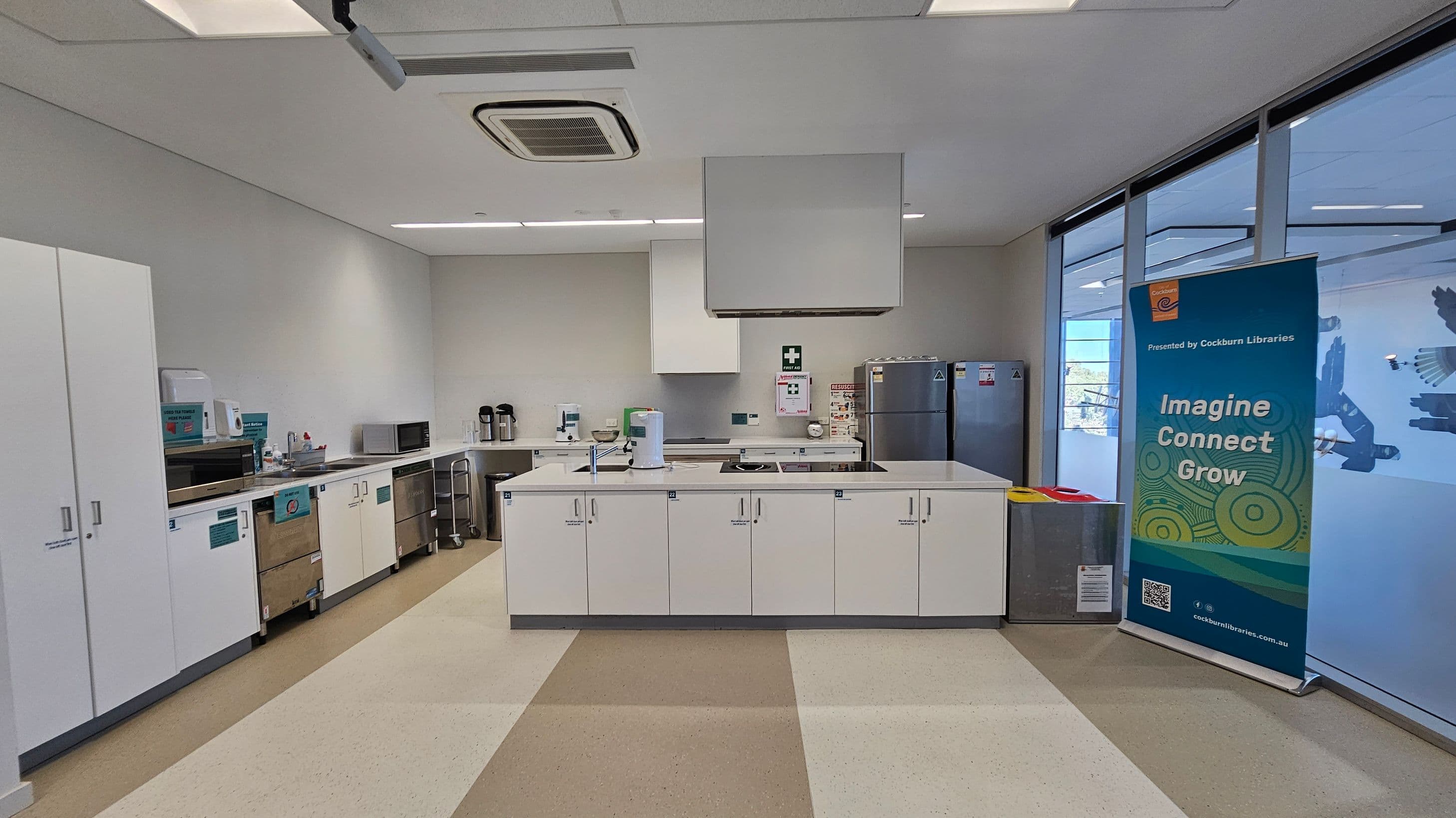 A modern kitchen area with white cabinetry and stainless steel countertops. On the left, there are cupboards, a microwave, and a sink with a drying rack. In the centre, a large island with a stovetop and a paper towel holder. On the right, a refrigerator and a blue banner with the text "Imagine Connect Grow" from Cockburn Libraries. The floor has a beige and brown checkered pattern, and the ceiling features recessed lighting and an air conditioning unit. Large windows are on the right.