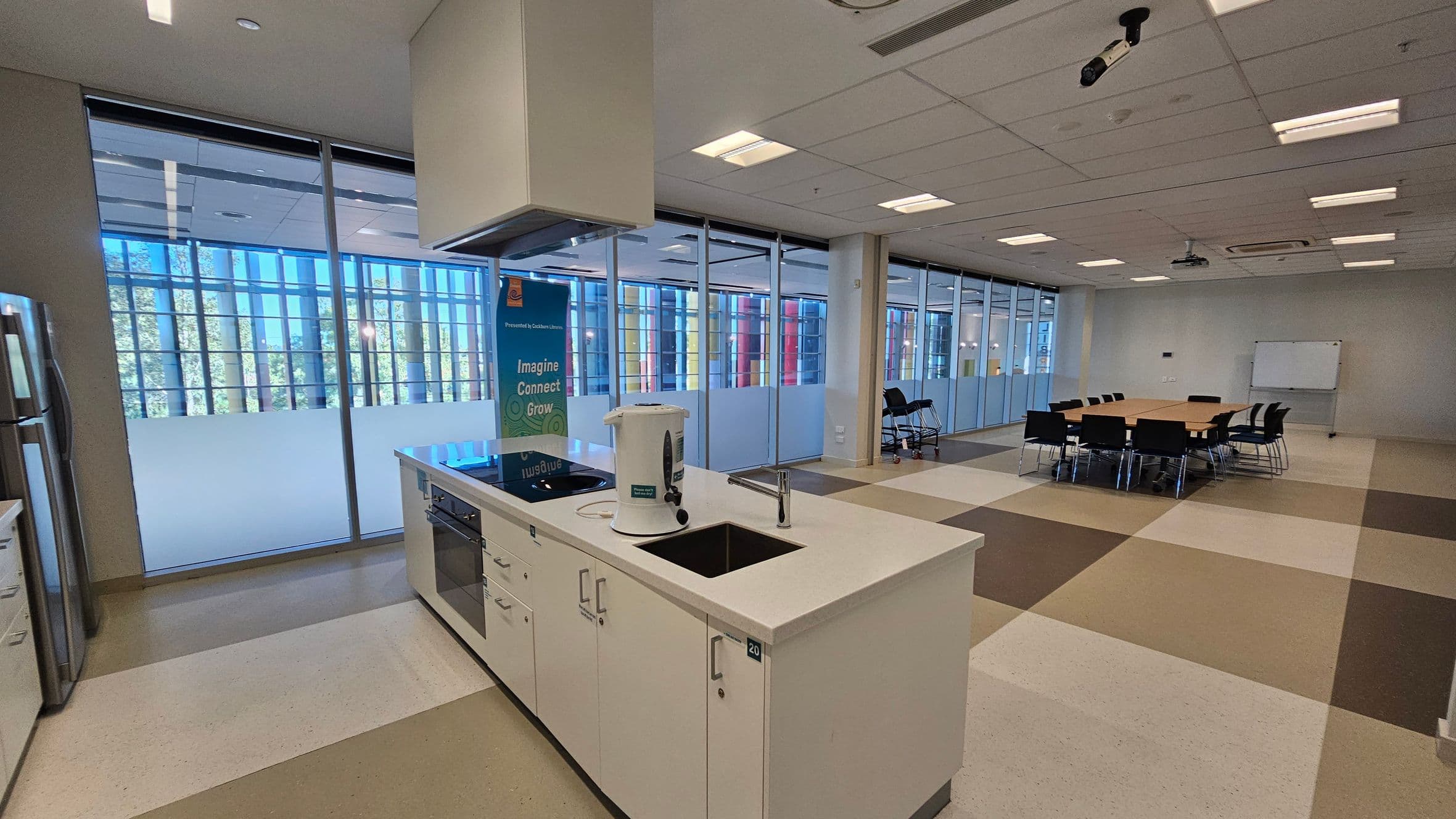 A modern office space with a kitchen area on the left, featuring a white countertop, sink, and a kettle. Behind it, large windows with frosted lower sections and colourful vertical blinds allow natural light. On the right, a meeting area with a rectangular table surrounded by black chairs. A whiteboard is visible against the far wall. The ceiling has recessed lighting and a projector. The floor is tiled in a checkerboard pattern of beige, brown, and white.
