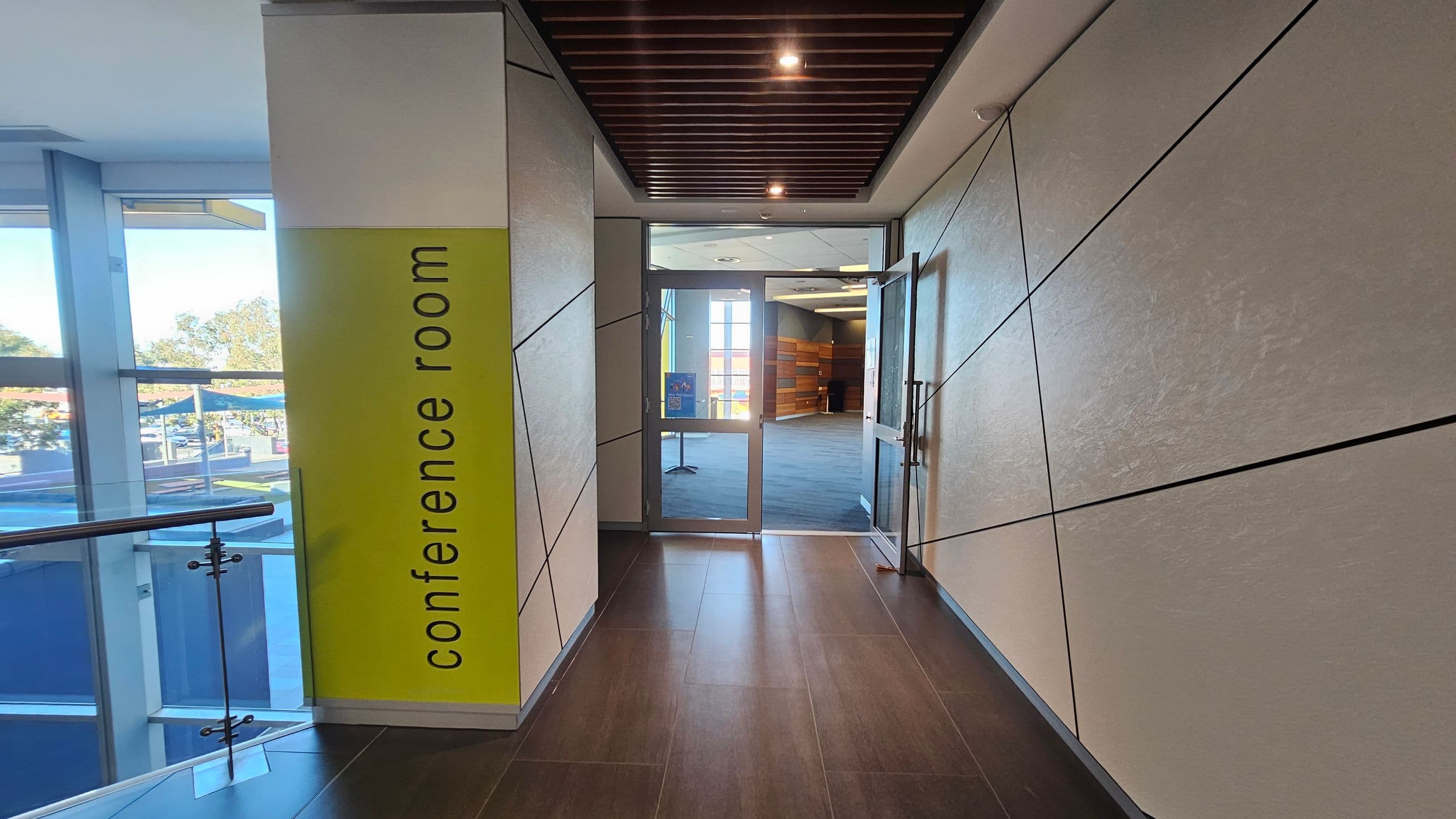 A hallway with a brown tiled floor and a ceiling featuring wooden slats and recessed lighting. On the left, a large window with a glass railing overlooks an outdoor area. A yellow wall with "conference room" written vertically is also on the left. The right wall is lined with light-coloured panels intersected by dark lines. At the end of the hallway, glass doors lead into a carpeted conference room with wooden wall accents and large windows.