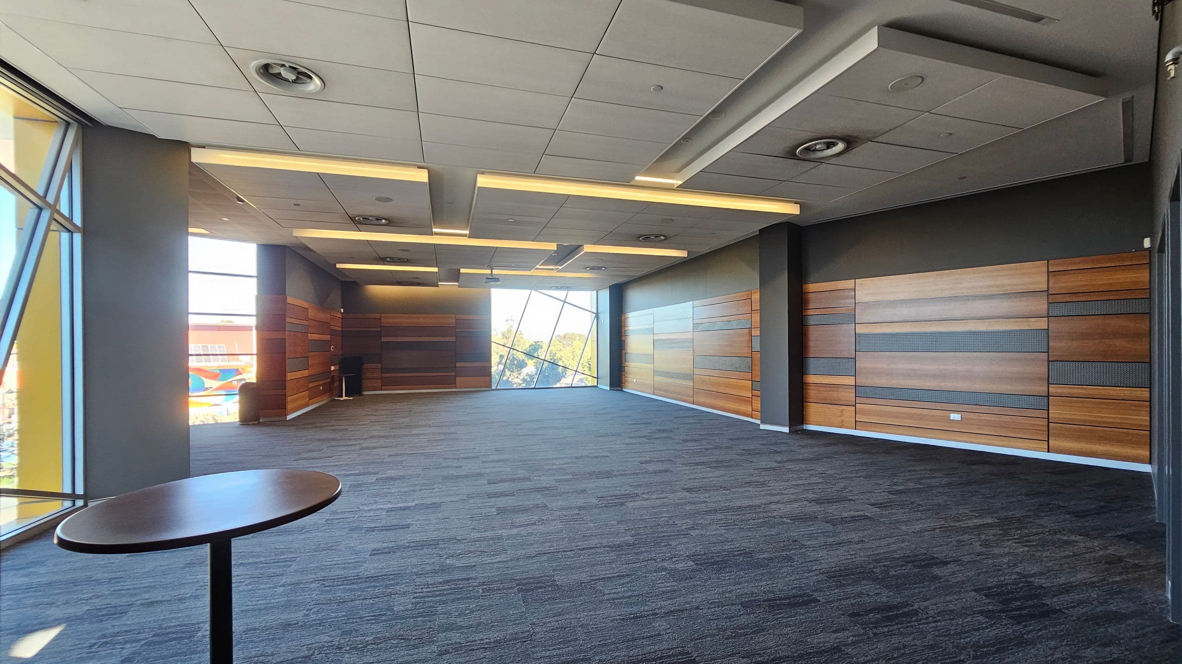 A spacious room with a modern design. The left side features large windows with a view of a colourful exterior. A small round table is near the window. The right side has wooden panelled walls with horizontal stripes. The ceiling is fitted with recessed lighting and ventilation fixtures. The floor is carpeted in a mix of grey and purple tones. The back wall has a large, slanted window offering a view of trees outside.
